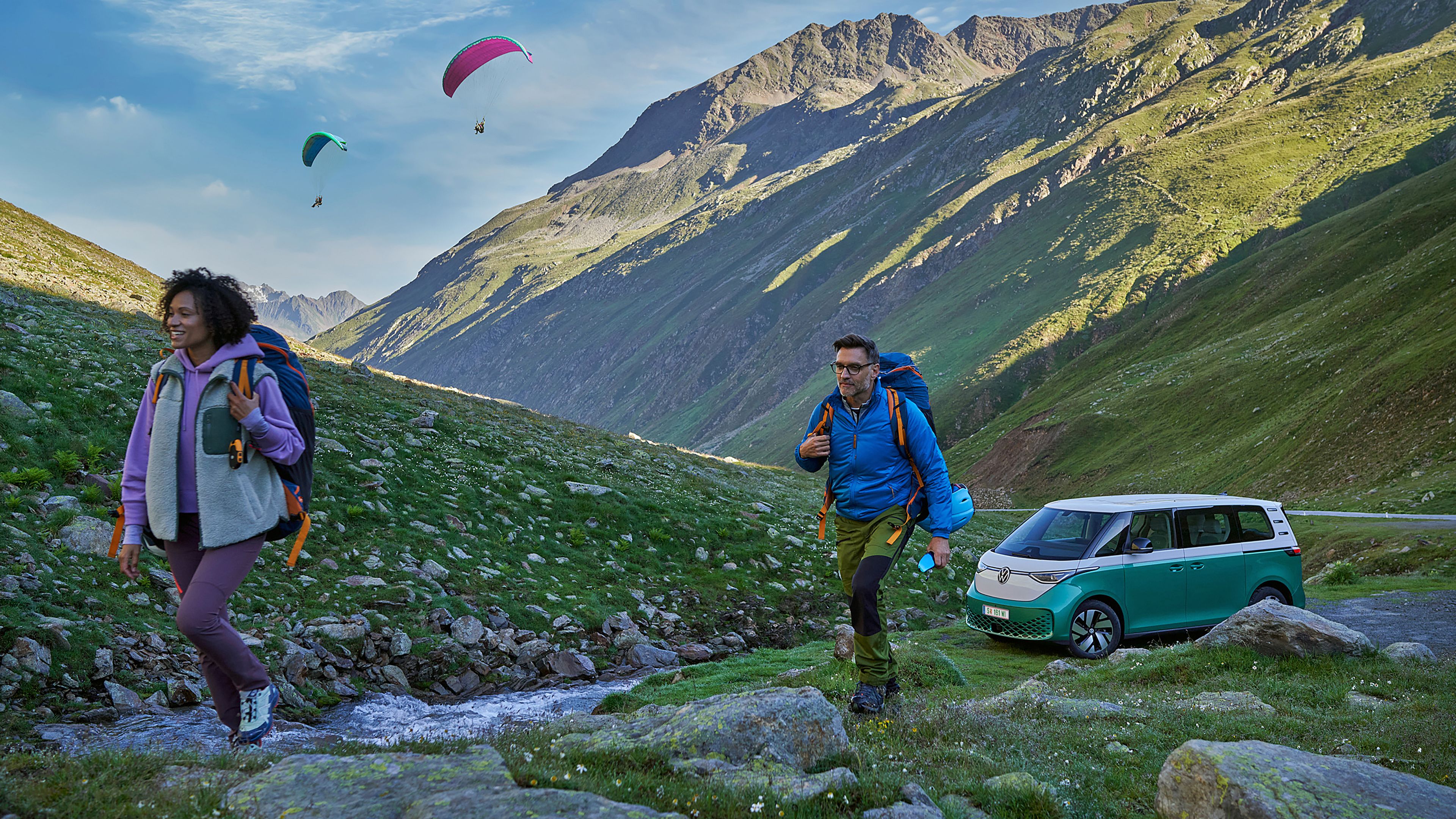 Двама туристи и електрически микробус Volkswagen ID. Buzz в живописен планински пейзаж с парапланери в небето