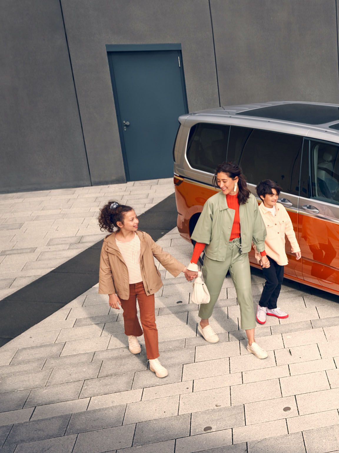 Щастливо семейство до оранжев Volkswagen Multivan eHybrid, паркиран до зарядна станция.
