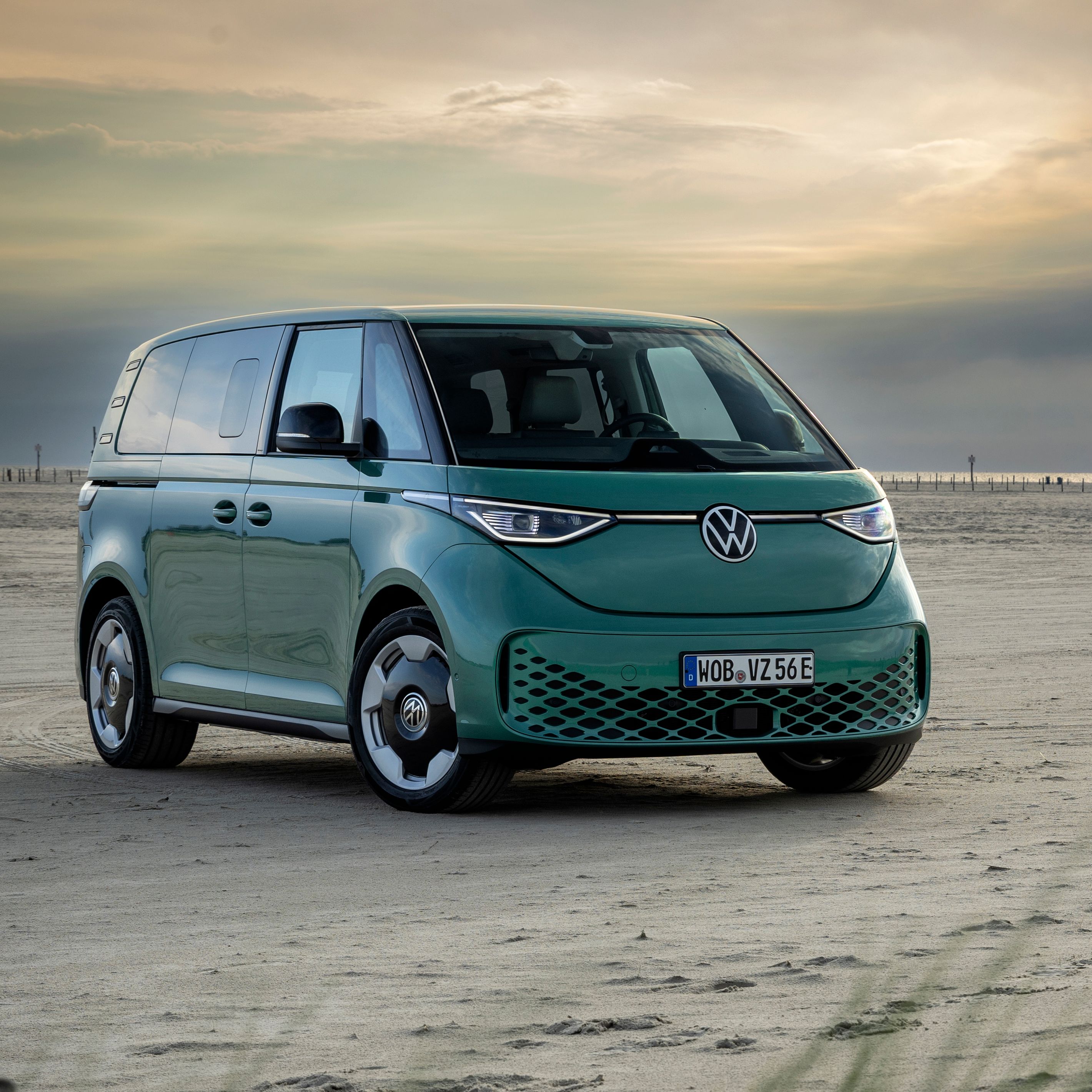Зелен микробус Volkswagen е паркиран на широк пуст плаж при залез слънце. В далечината се виждат морето и силует на постройка върху пилони, а небето е облачно с ярко отблясъчно петно от слънцето. На преден план има туфи зелена трева и дървени колове в пясъка.