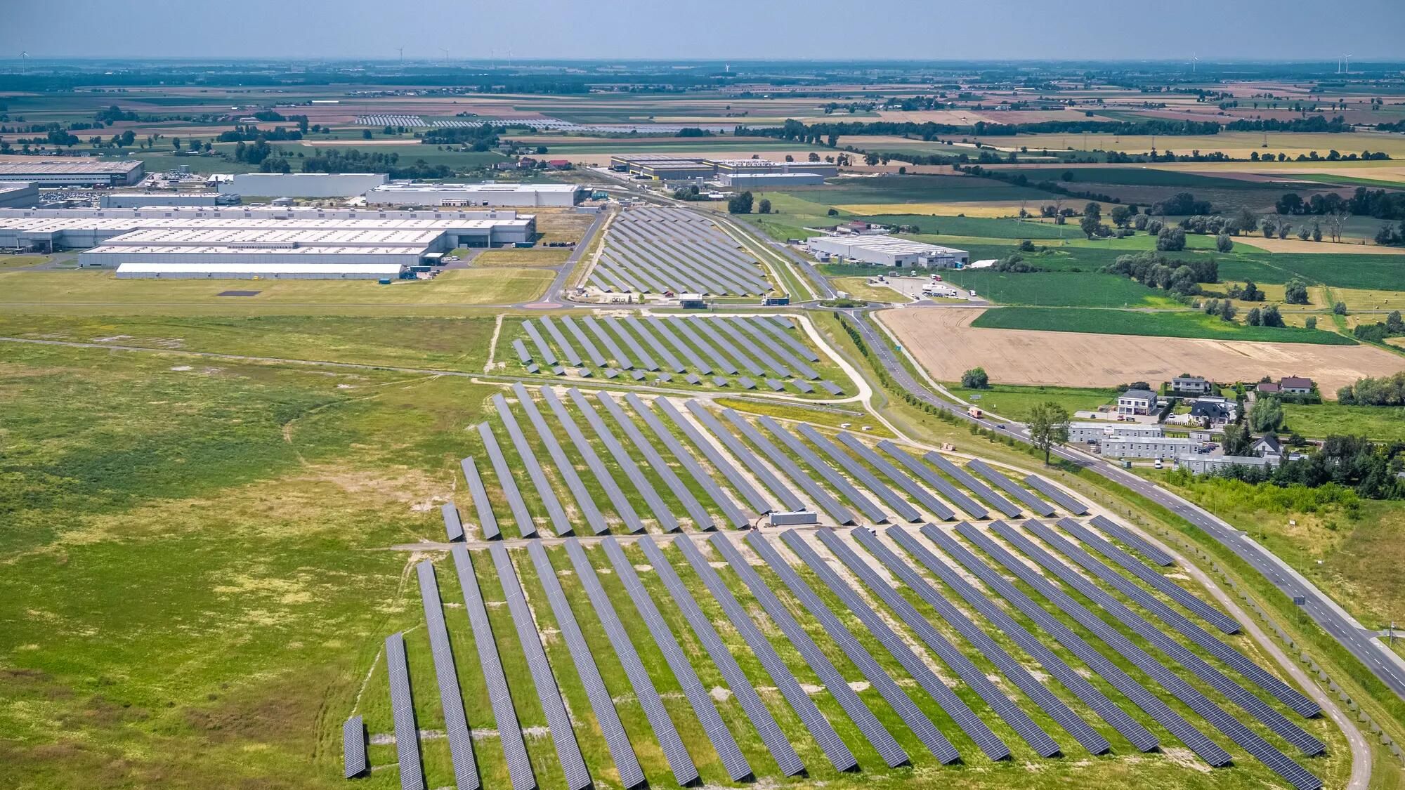 Соларен парк, разположен в селскостопанска зона във Вжешня, с много соларни панели, подредени в редове. Отзад се виждат промишлени сгради и обработваеми земи. Панорамата показва отдалечените полета и зелените площи наоколо.