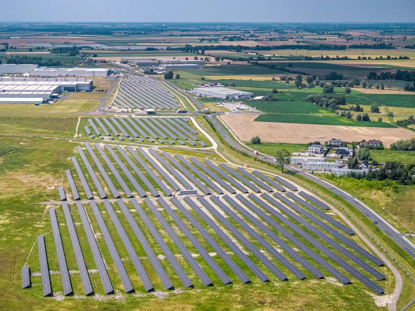 Соларен парк, разположен в селскостопанска зона във Вжешня, с много соларни панели, подредени в редове. Отзад се виждат промишлени сгради и обработваеми земи. Панорамата показва отдалечените полета и зелените площи наоколо.