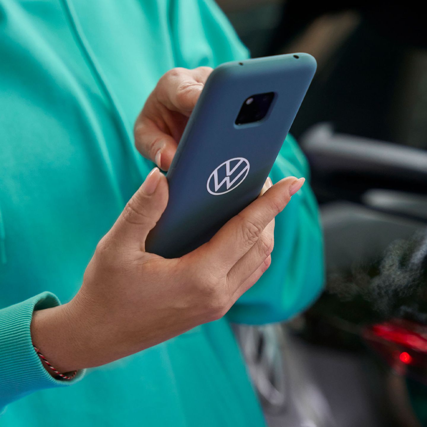 Човек, облечен в зелен суитчър, държи смартфон със знак на Volkswagen на гърба му. На преден план се вижда задната светлина на автомобил.