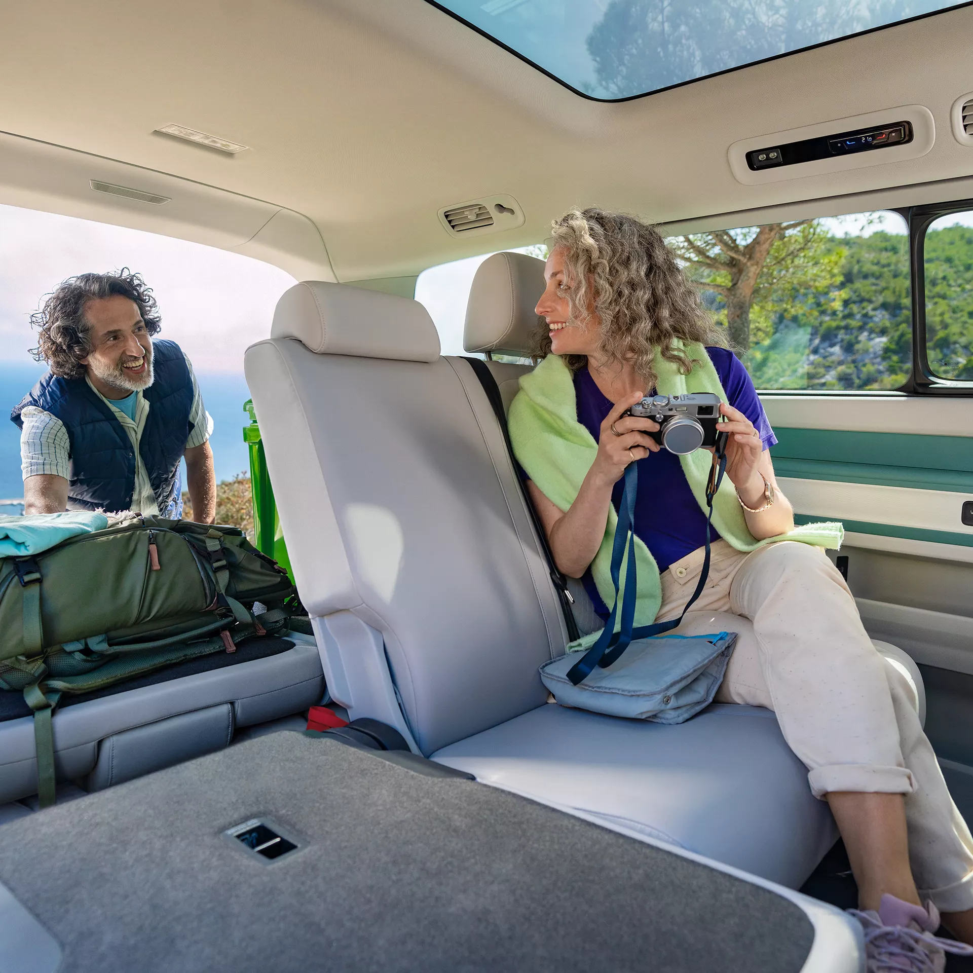 Двойка се наслаждава на почивка сред природата във вътрешността на Volkswagen ID. Buzz, с отворен багажник и фотоапарат в ръка.