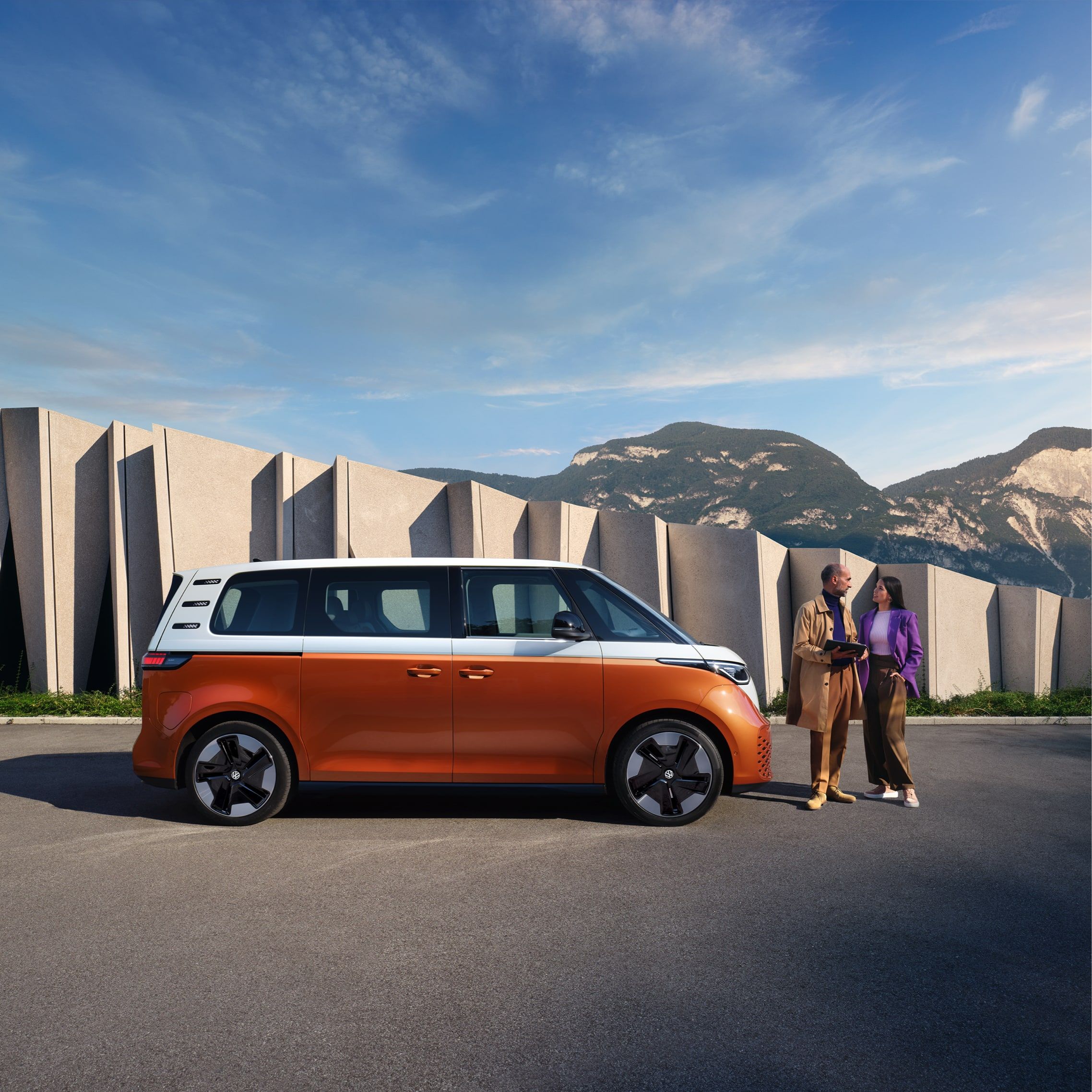 Volkswagen ID. Buzz паркиран пред модерна сграда и планински пейзаж с двойка в разговор