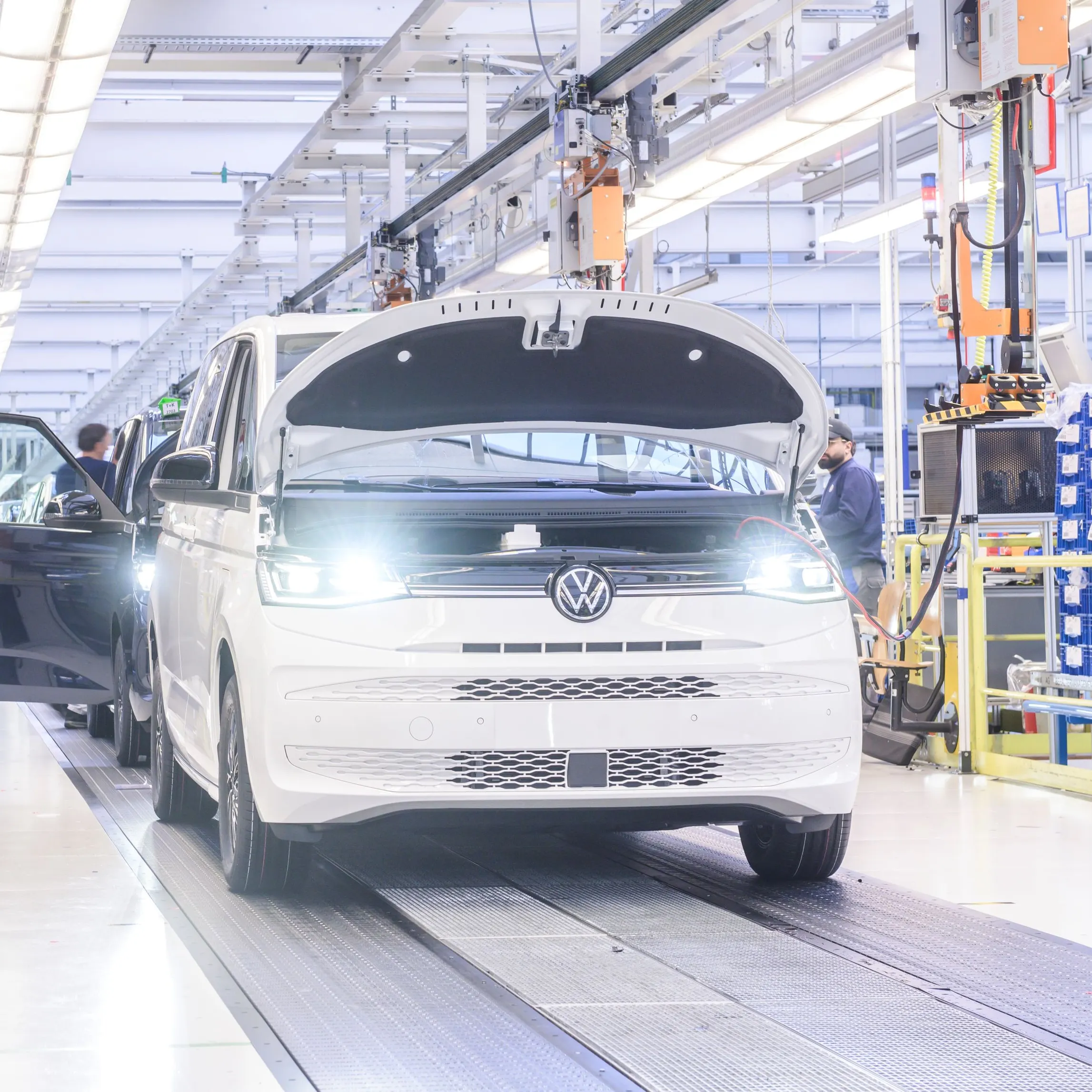Снимка на производствена линия във фабрика в Хановер, където се сглобяват бели микробуси Volkswagen. Микробусът на преден план има отворен капак, а по-нататък се виждат работници, занимаващи се с монтажа. Средата е добре осветена с промишлени светлини.