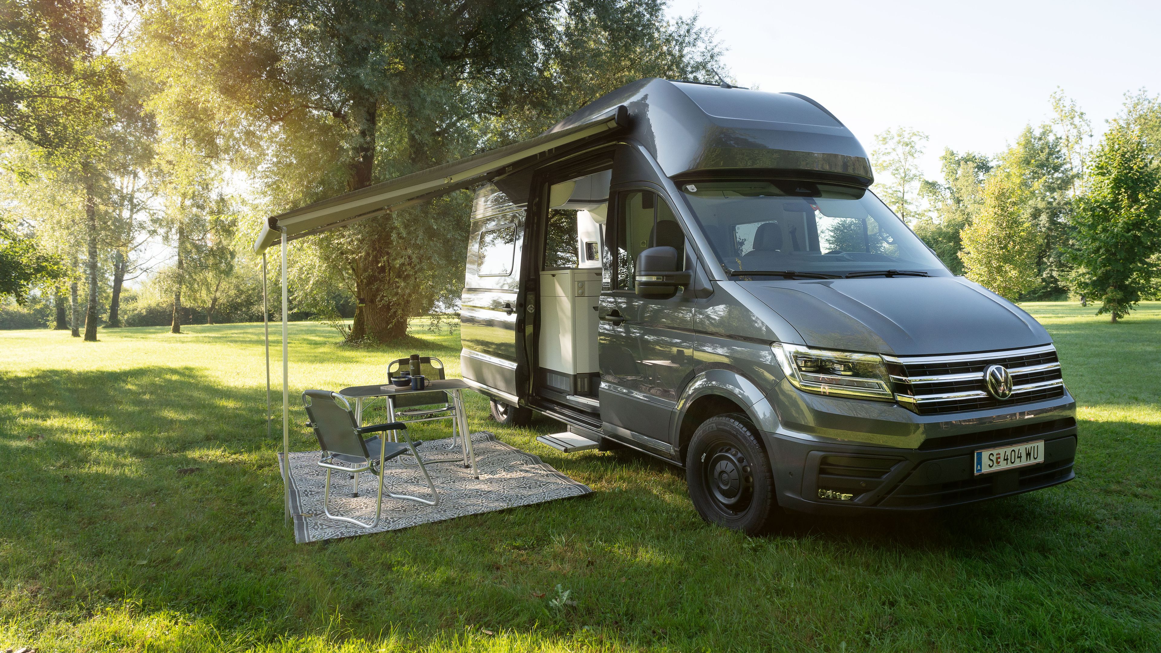 Ein grauer Campingbus steht auf einer grünen Wiese im Sonnenschein. Daneben sind Campingmöbel und ein Grill unter einer ausgefahrenen Markise platziert. Im Hintergrund sind Bäume und Sträucher zu sehen.