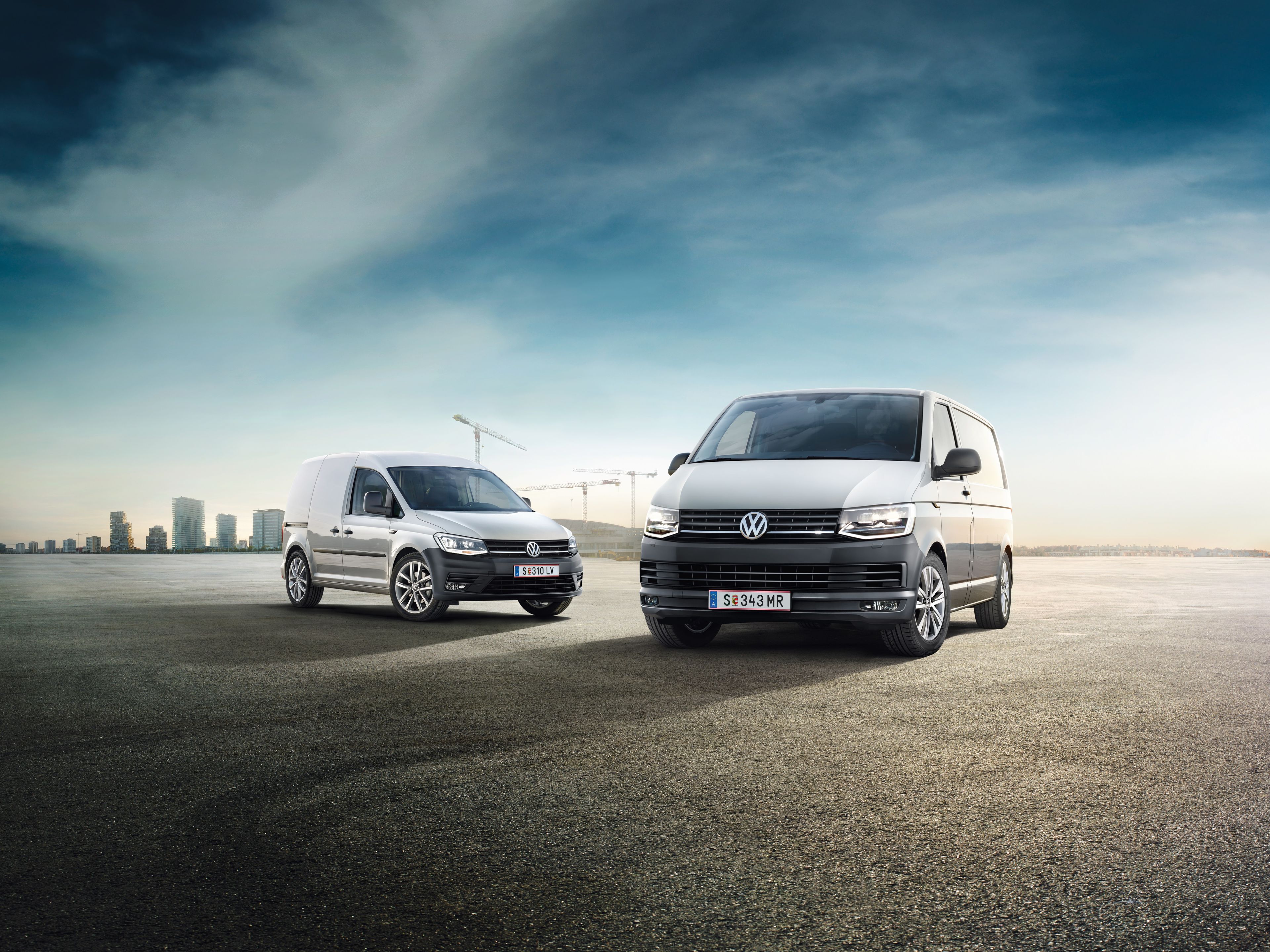 Два лекотоварни автомобила Volkswagen са паркирани на голямо открито пространство, като на заден план се вижда градски пейзаж с високи сгради.