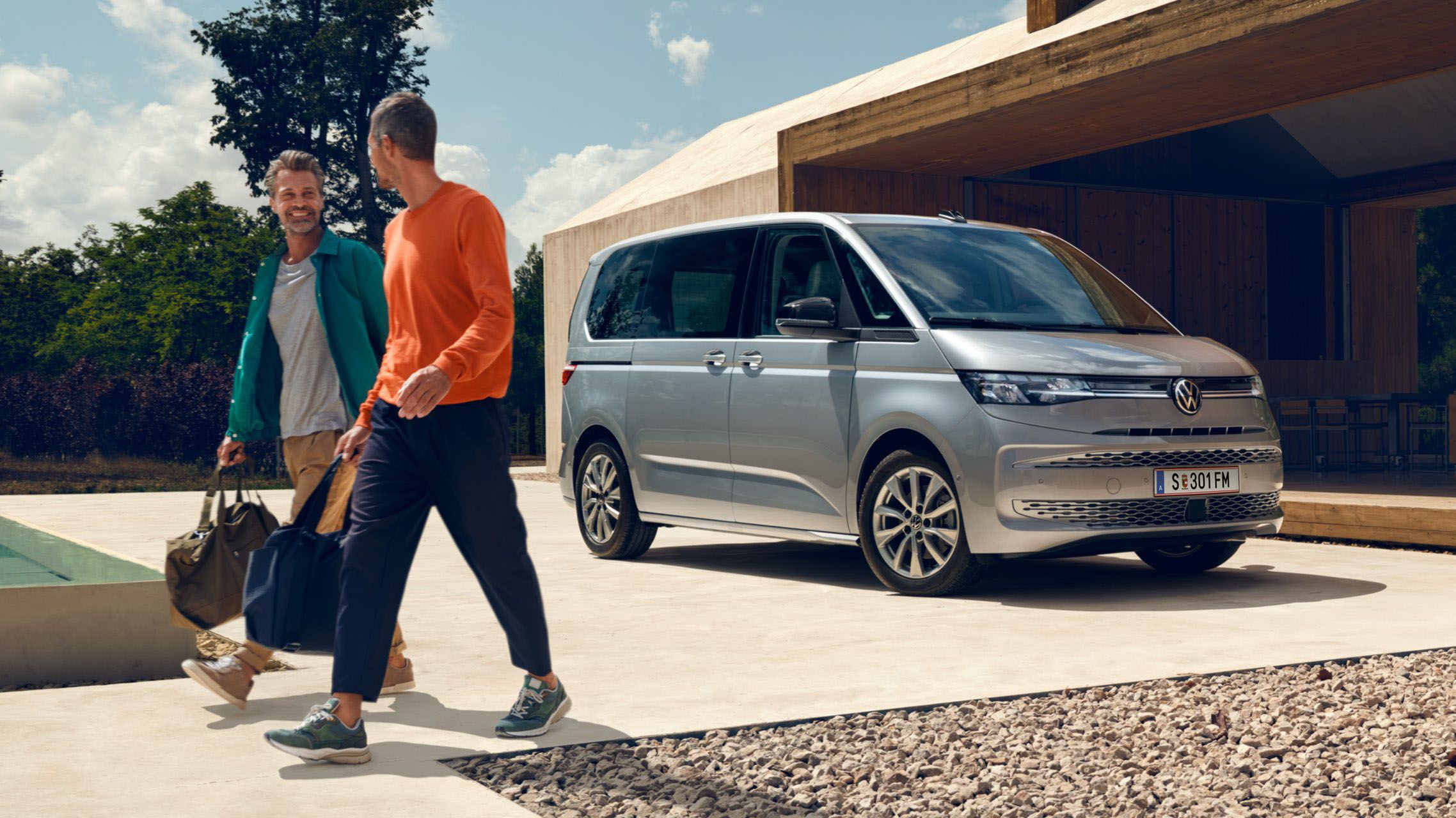 Zwei Männer mit Sporttaschen laufen vor einem neuen silbernen VW Multivan T7 2021.