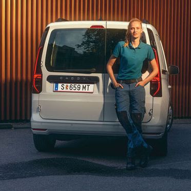 Жена, облечена в работно облекло, облягаща се на задната част на Volkswagen Caddy, паркиран пред модерна фасада.