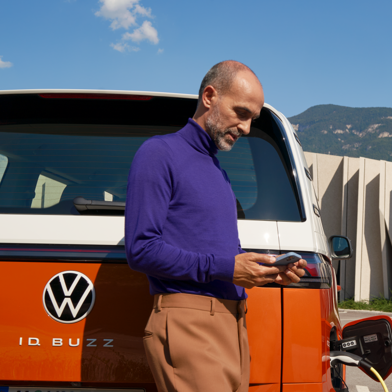 Мъж зарежда електрическия Volkswagen ID. Buzz на зарядна станция, докато проверява телефона си.