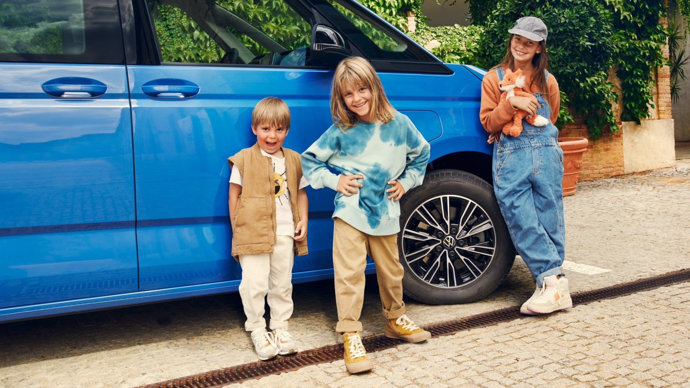 Drei Kinder stehen vor einem blauen neuen VW Multivan T7 2021.
