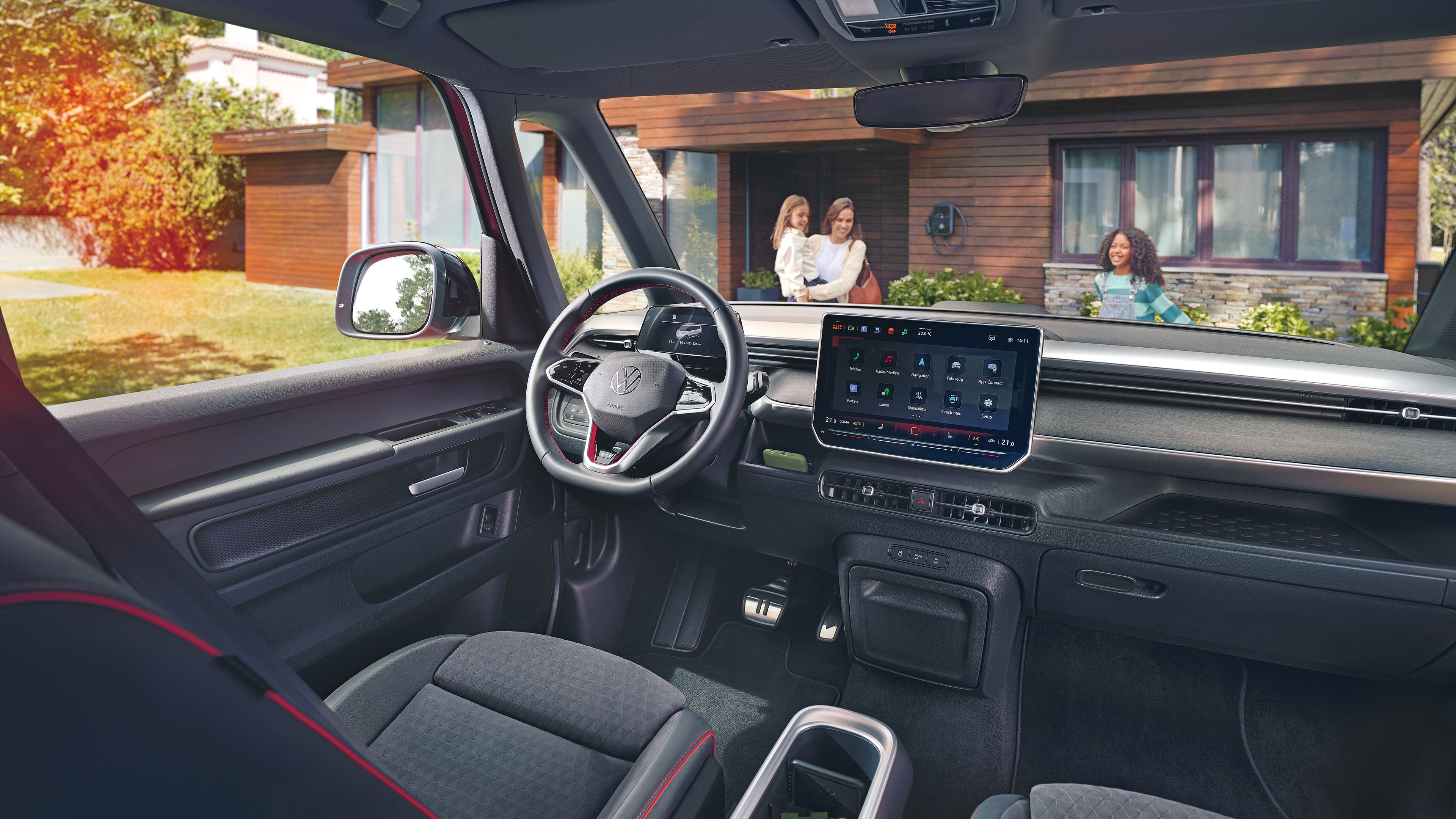 Интериор на Volkswagen ID. Buzz с модерен дигитален кокпит и изглед към дом с три жени пред зарядна станция.