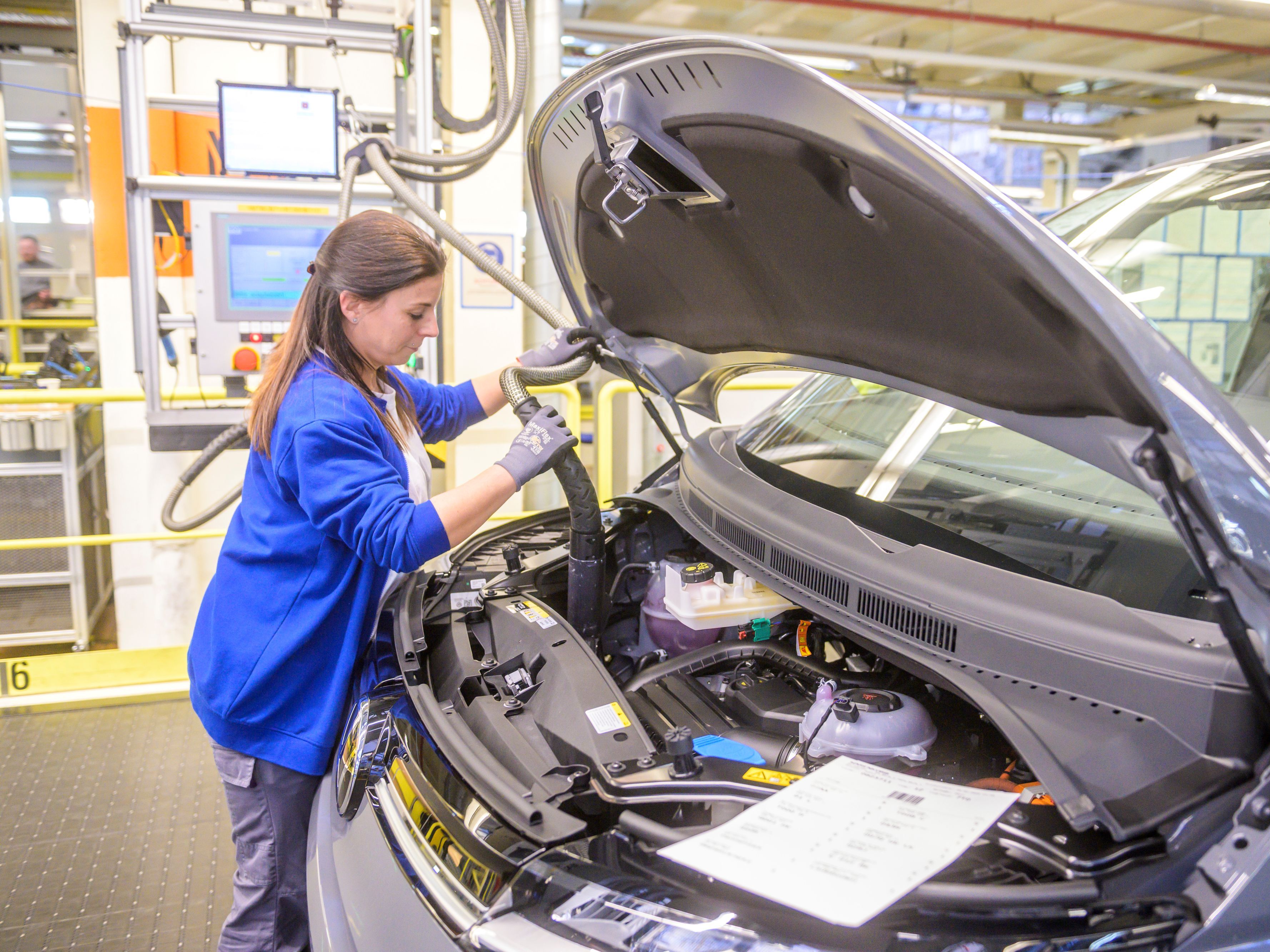 Жена в синя работна риза инспектира моторния отсек на Volkswagen автомобил в производствено хале. Капакът на автомобила е повдигнат, а около нея има индустриално оборудване и компютърни екрани.