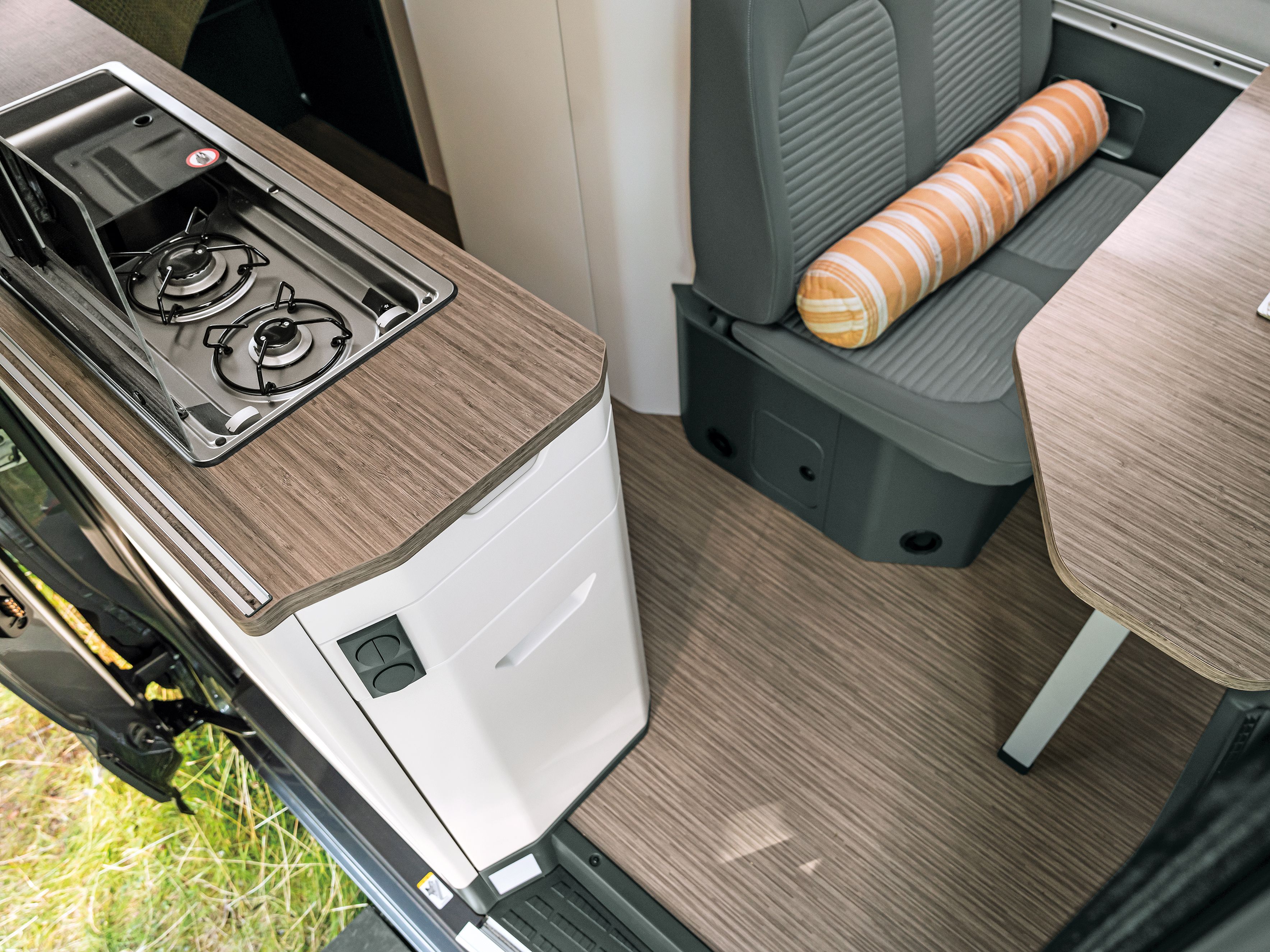 Innenansicht eines Wohnmobils mit einem Gaskocher, einer kleinen Arbeitsfläche und einem Sitzbereich mit einem Tisch. Auf dem Tisch steht eine kleine Vase mit gelben Blumen. Der Sitzbereich ist grau und hat ein gestreiftes Kissen.