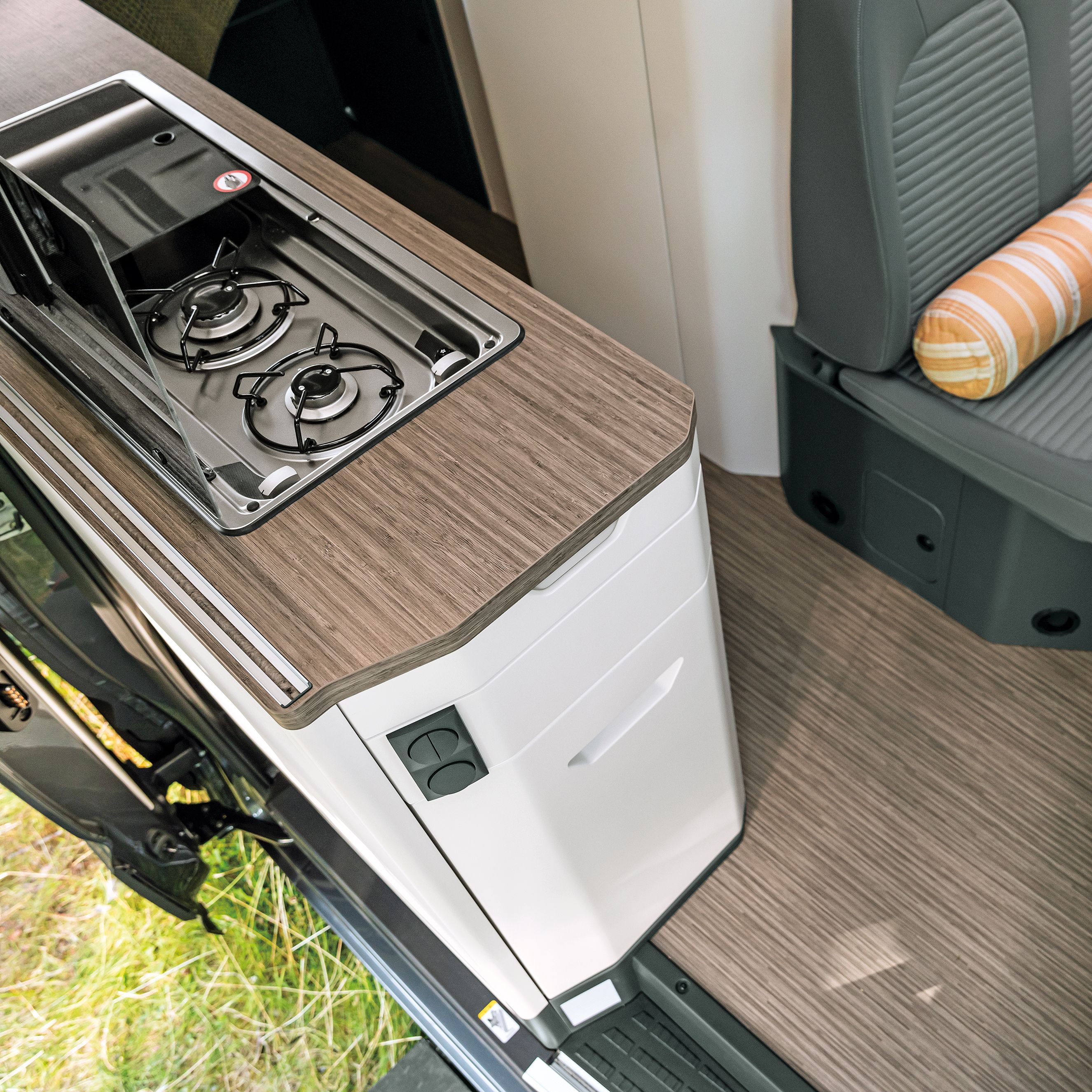 Innenansicht eines Wohnmobils mit einem Gaskocher, einer kleinen Arbeitsfläche und einem Sitzbereich mit einem Tisch. Auf dem Tisch steht eine kleine Vase mit gelben Blumen. Der Sitzbereich ist grau und hat ein gestreiftes Kissen.