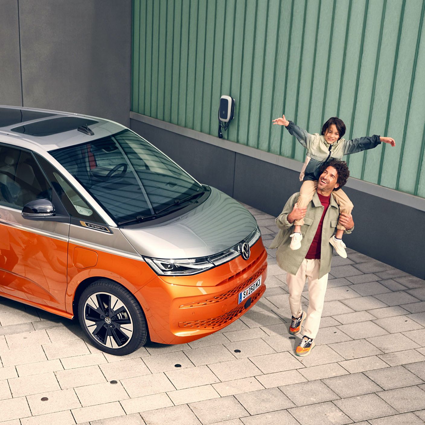 Щастливо семейство до оранжев Volkswagen Multivan eHybrid, паркиран до зарядна станция.
