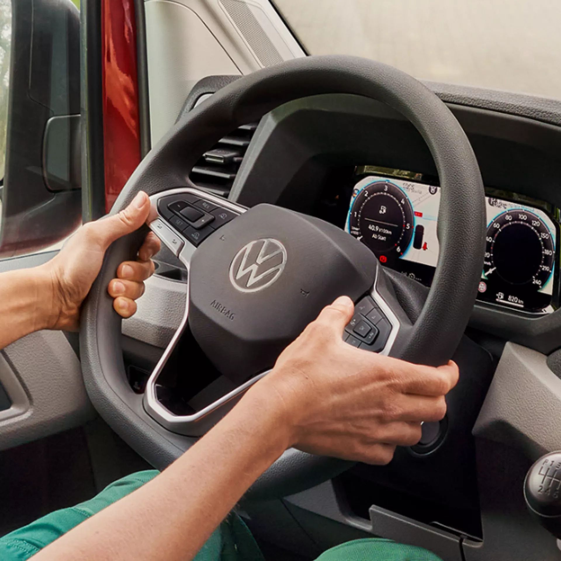 Човек държи волана на автомобил, на който се вижда логото на Volkswagen. На таблото пред него има два цифрови дисплея. Ръцете му са разположени в позиция "девет и три".