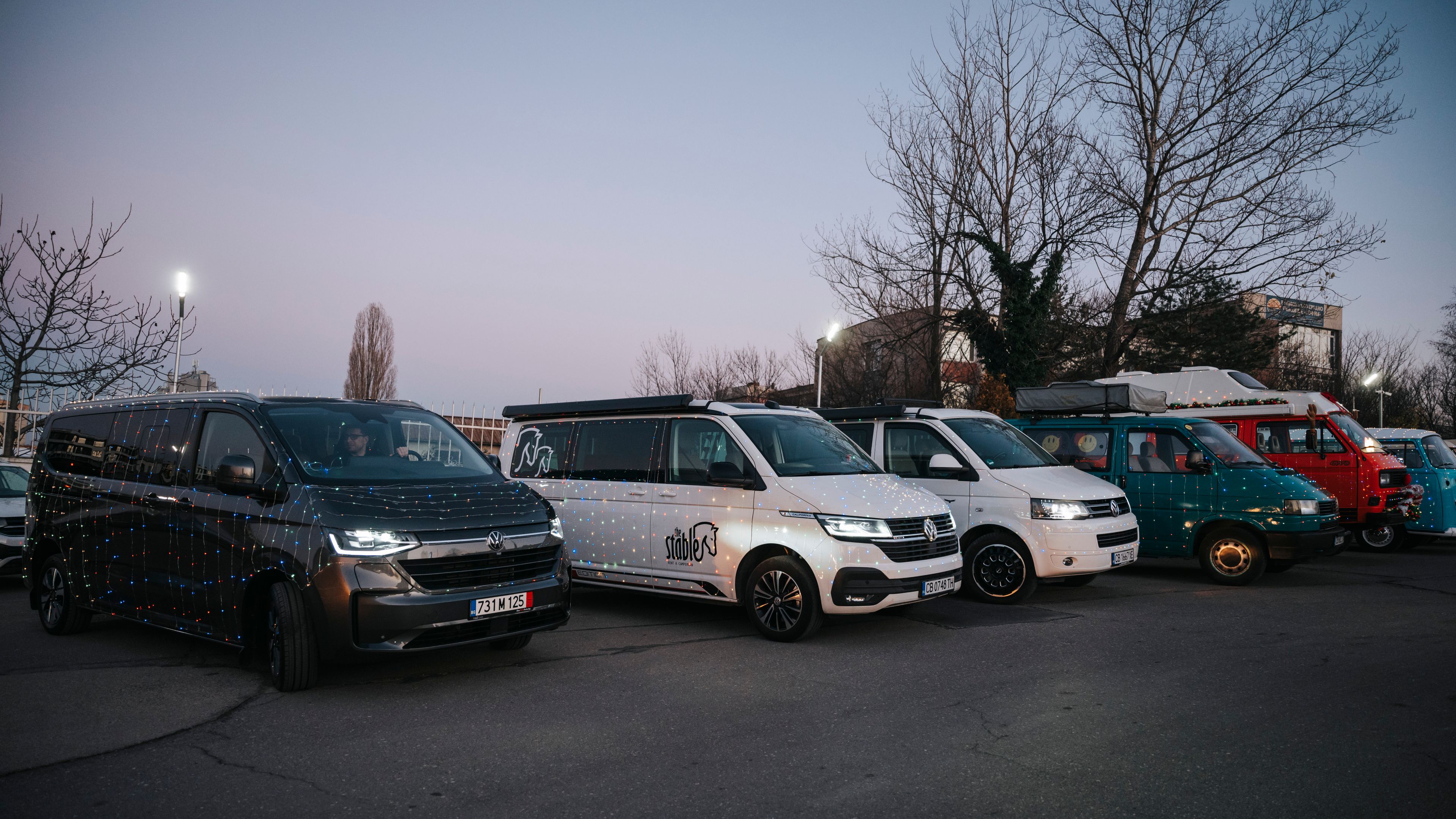 Снимката показва редица паркирани Volkswagen микробуси на открит паркинг. Зад микробусите има дървета без листа. Небето е ясно и леко осветено от залеза.