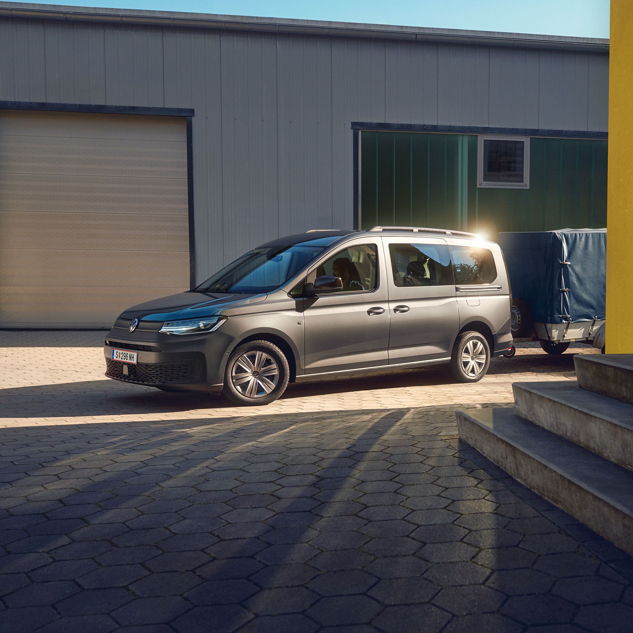 Сив Volkswagen Caddy с прикачено ремарке, паркиран пред индустриална сграда с жълта фасада, на залез слънце.