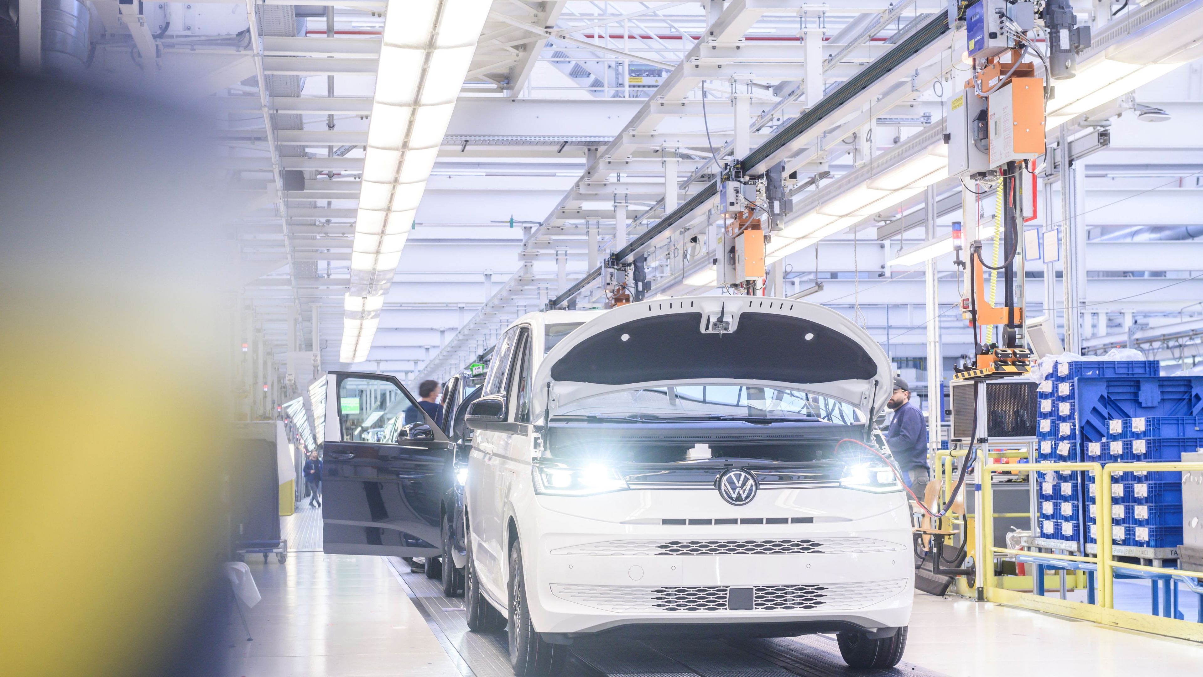 Снимка на производствена линия във фабрика в Хановер, където се сглобяват бели микробуси Volkswagen. Микробусът на преден план има отворен капак, а по-нататък се виждат работници, занимаващи се с монтажа. Средата е добре осветена с промишлени светлини.