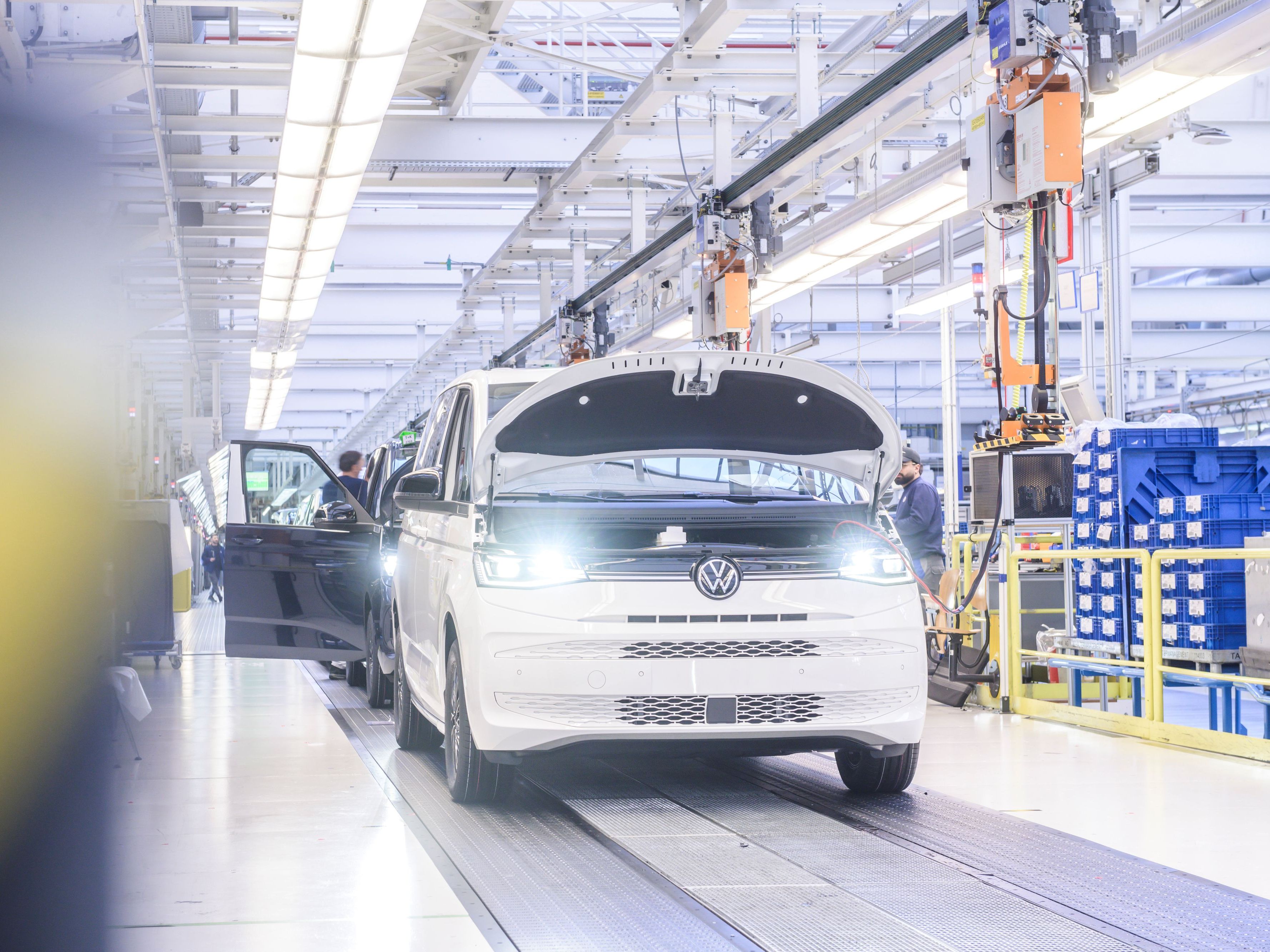 Снимка на производствена линия във фабрика в Хановер, където се сглобяват бели микробуси Volkswagen. Микробусът на преден план има отворен капак, а по-нататък се виждат работници, занимаващи се с монтажа. Средата е добре осветена с промишлени светлини.