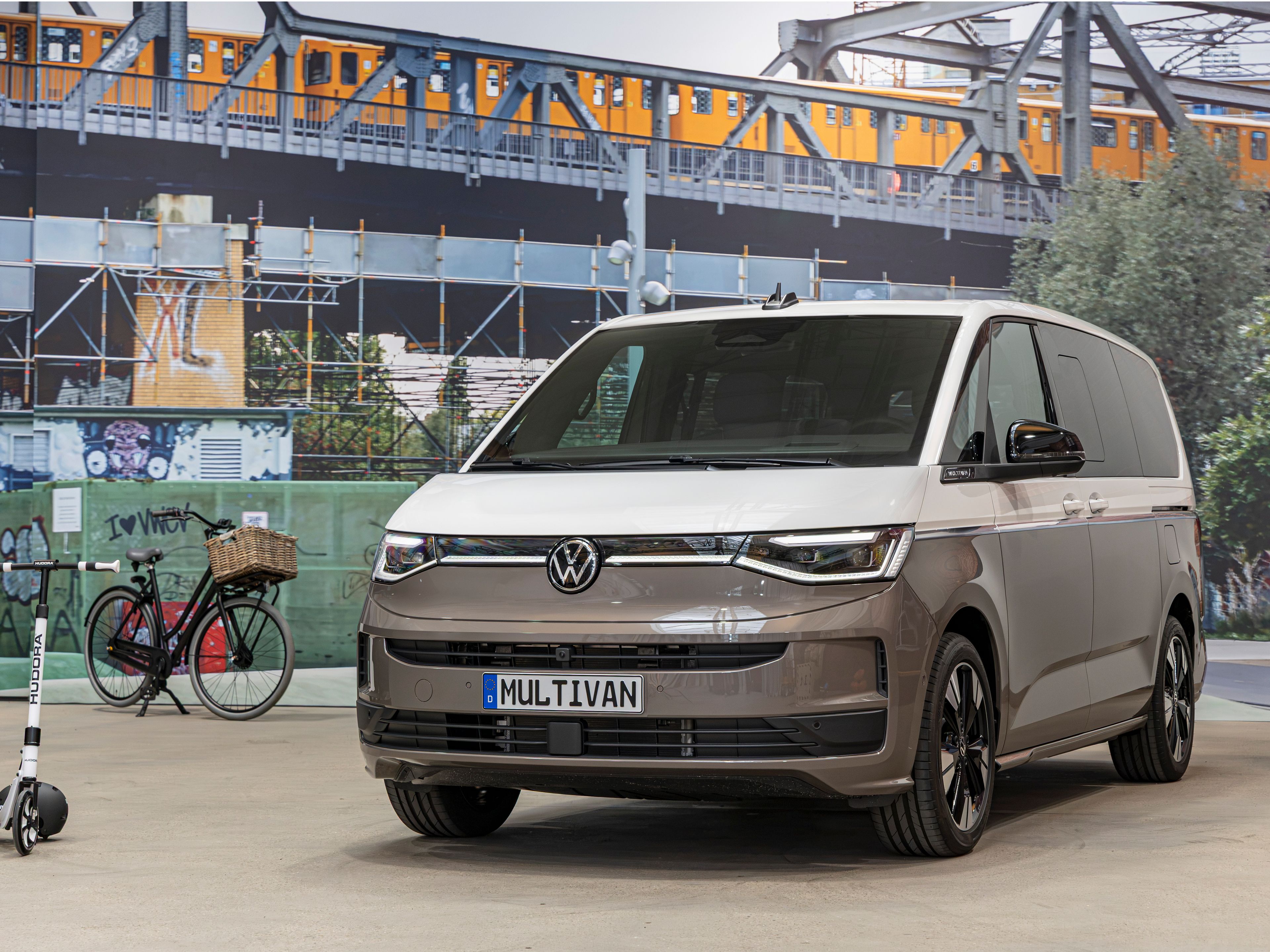 Съвременен миниван Volkswagen Multivan в бежово-сив цвят е паркиран на закрито пред голям стенопис със железопътен мост и преминаващ влак. Вляво има паркирано колело с кошница и тротинетка, оставени до стената. Атмосферата напомня градска среда с акцент върху модерна мобилност.