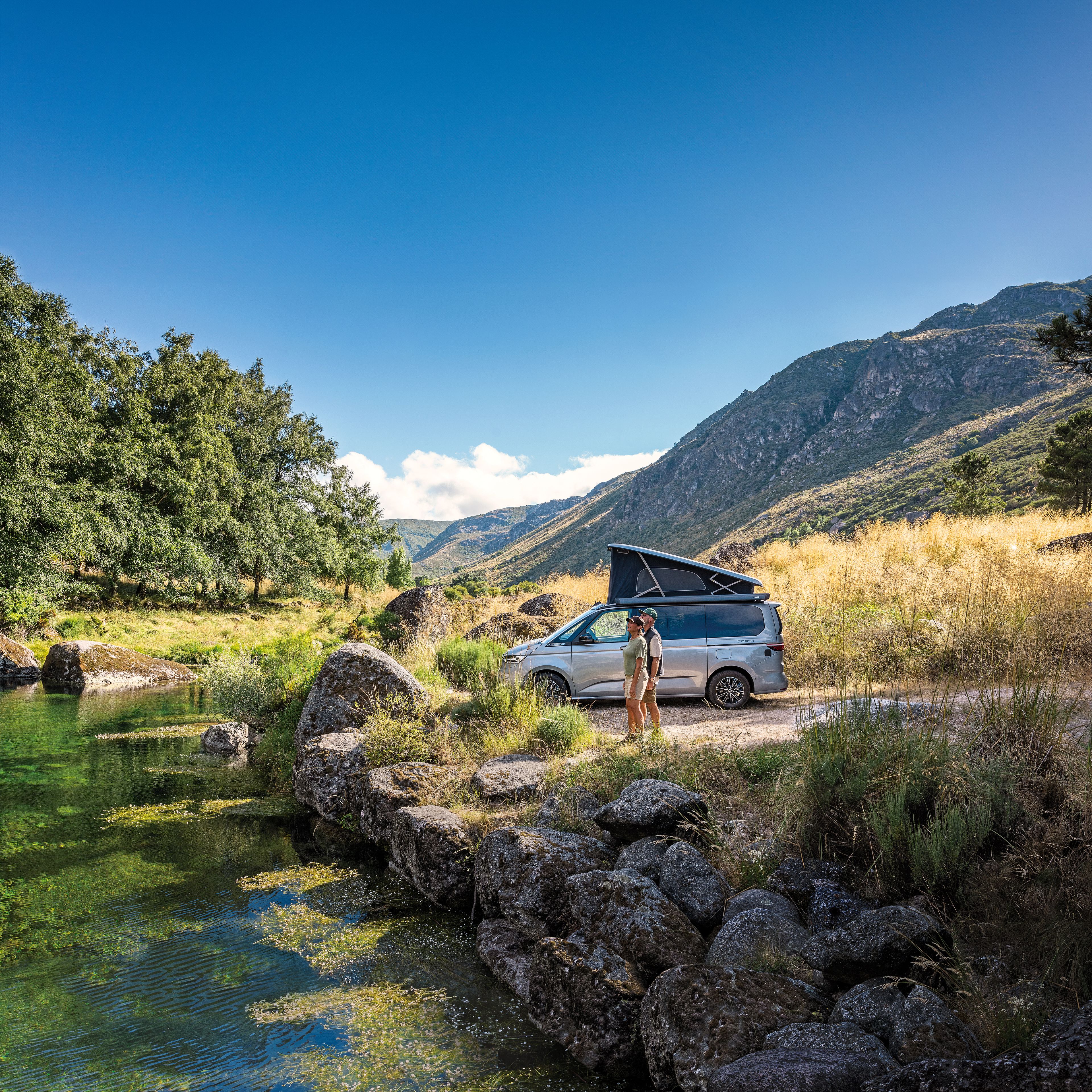 Двойка с кемпер Volkswagen, паркиран край планинско езеро сред живописна природа.