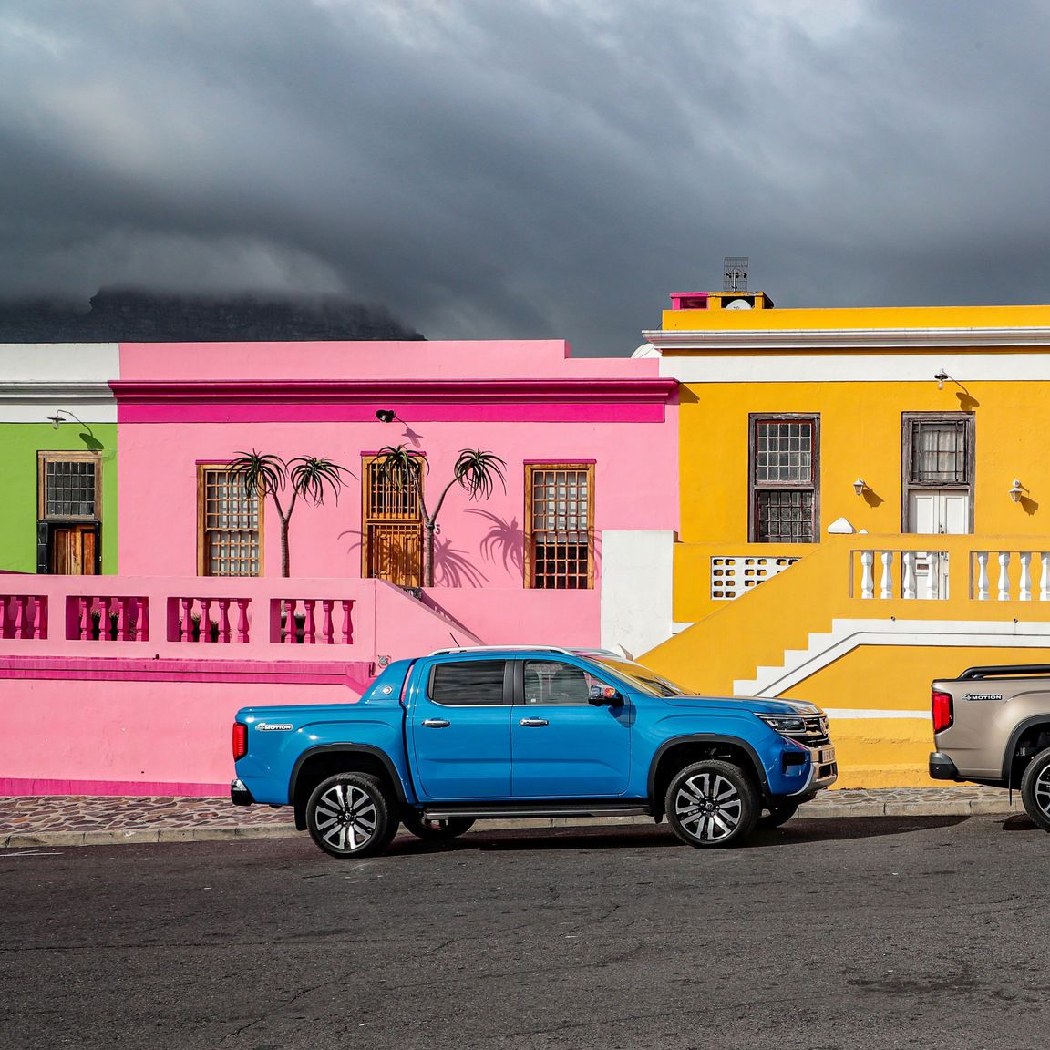 Два пикапа Volkswagen, паркирани на улица пред къщи в ярки цветове – розово, жълто и червено.