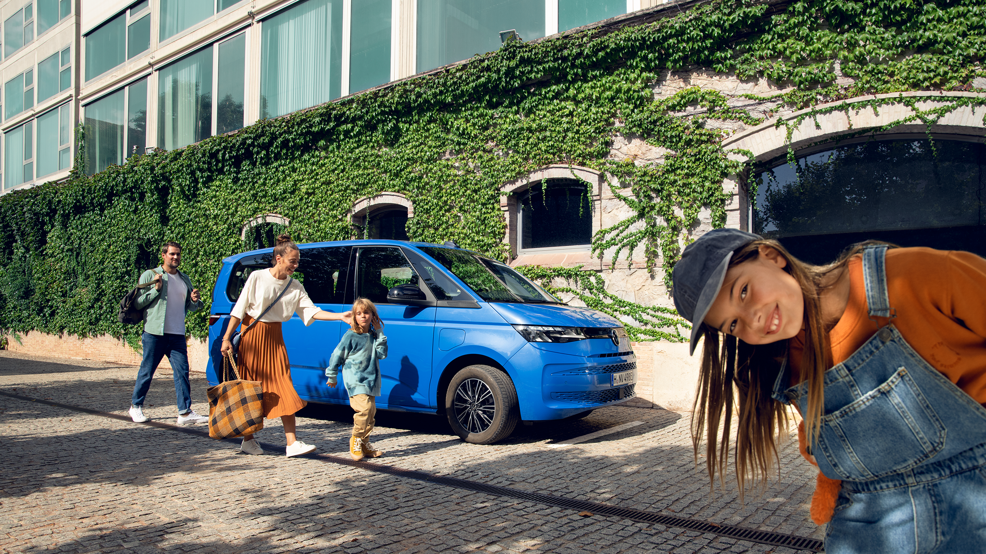 Семейно пътуване със стил – Volkswagen Multivan Син Volkswagen Multivan, паркиран на улица с модерна сграда, семейство с деца излиза от автомобила