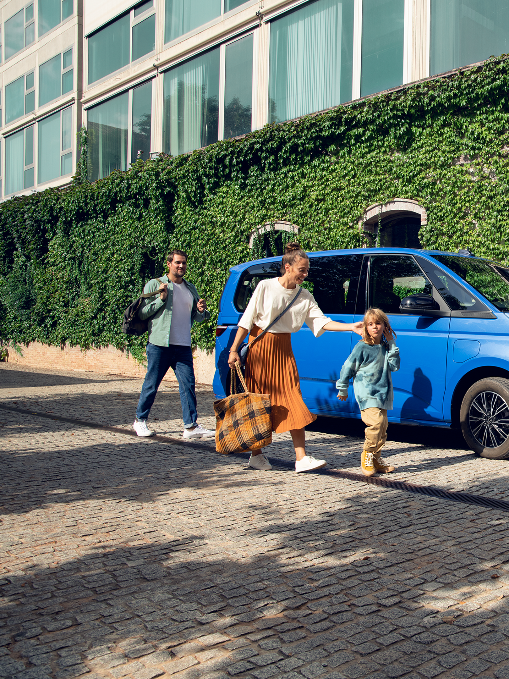 Семейно пътуване със стил – Volkswagen Multivan Син Volkswagen Multivan, паркиран на улица с модерна сграда, семейство с деца излиза от автомобила