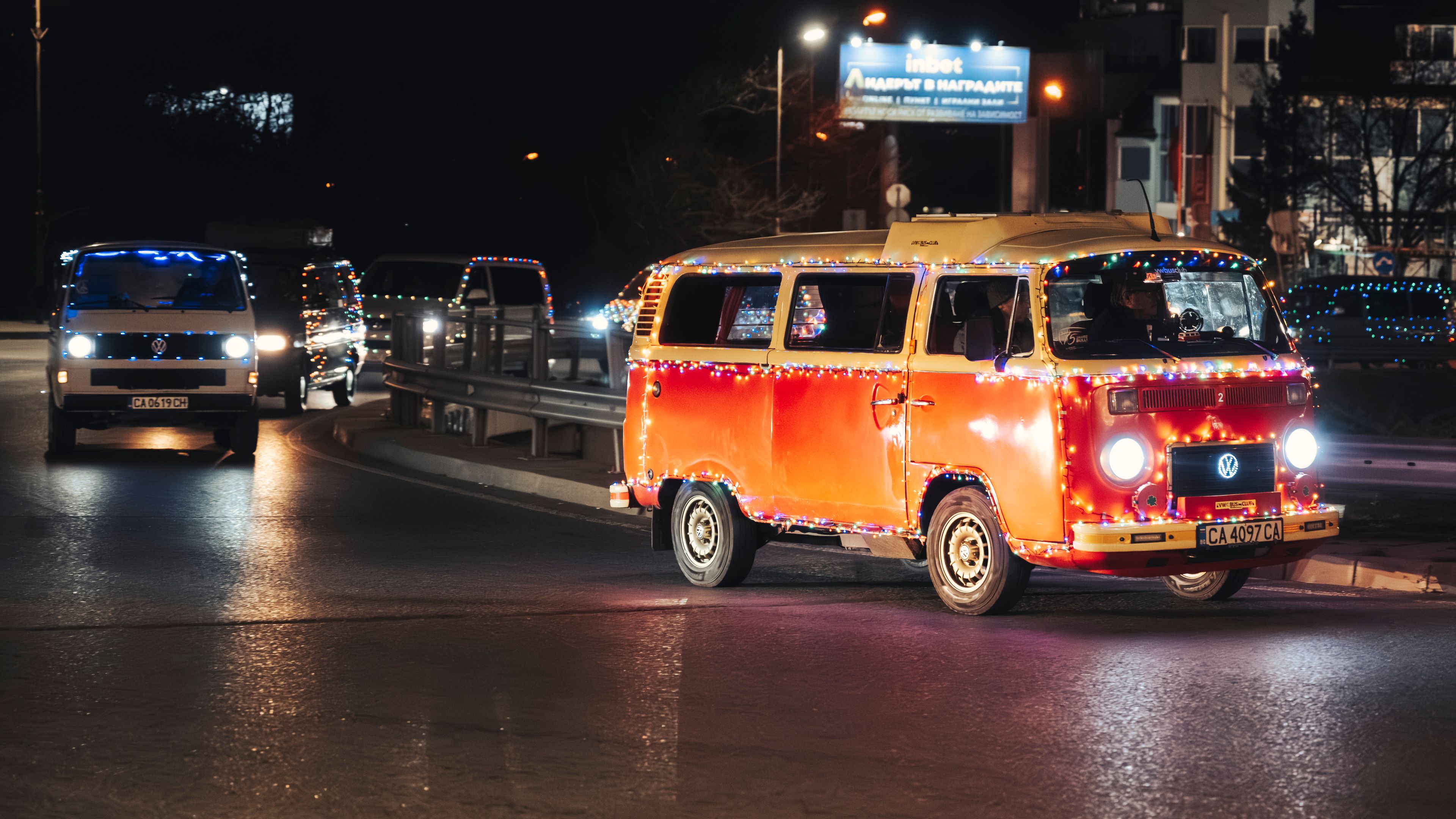 Ретро Volkswagen микробус, украсен със светлини, кара през нощта по осветен улица. На заден план се виждат други автомобили, както и светещи реклами. Сцената е празнична и оживена.