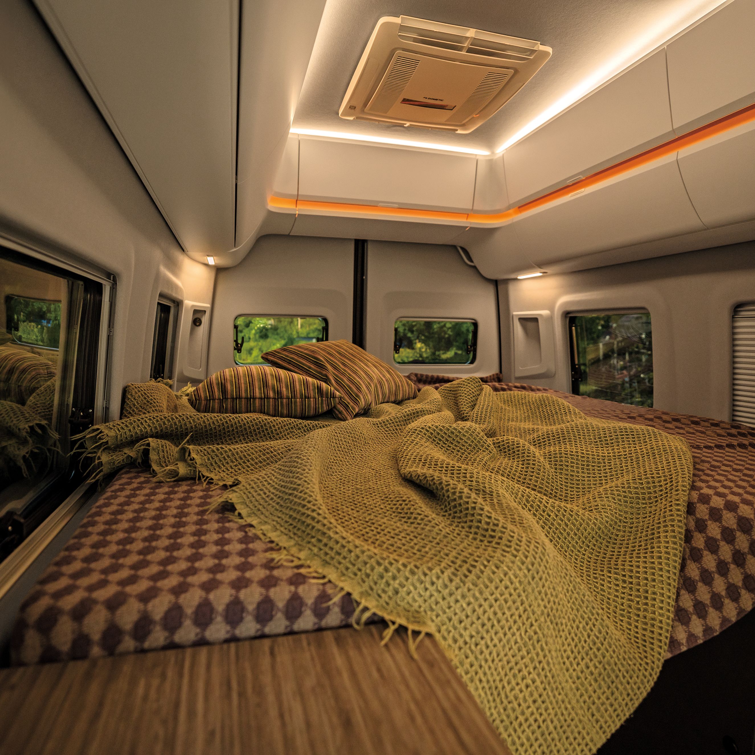 Das Bild zeigt das Interieur eines Wohnmobils mit einem gemütlichen Bett, das mit braun-gemusterten Bettlaken und gelblichen Decken bedeckt ist. An den Seiten sind Fenster zu sehen, durch die grüne Landschaft sichtbar ist. Oben an der Decke befinden sich Beleuchtungselemente.