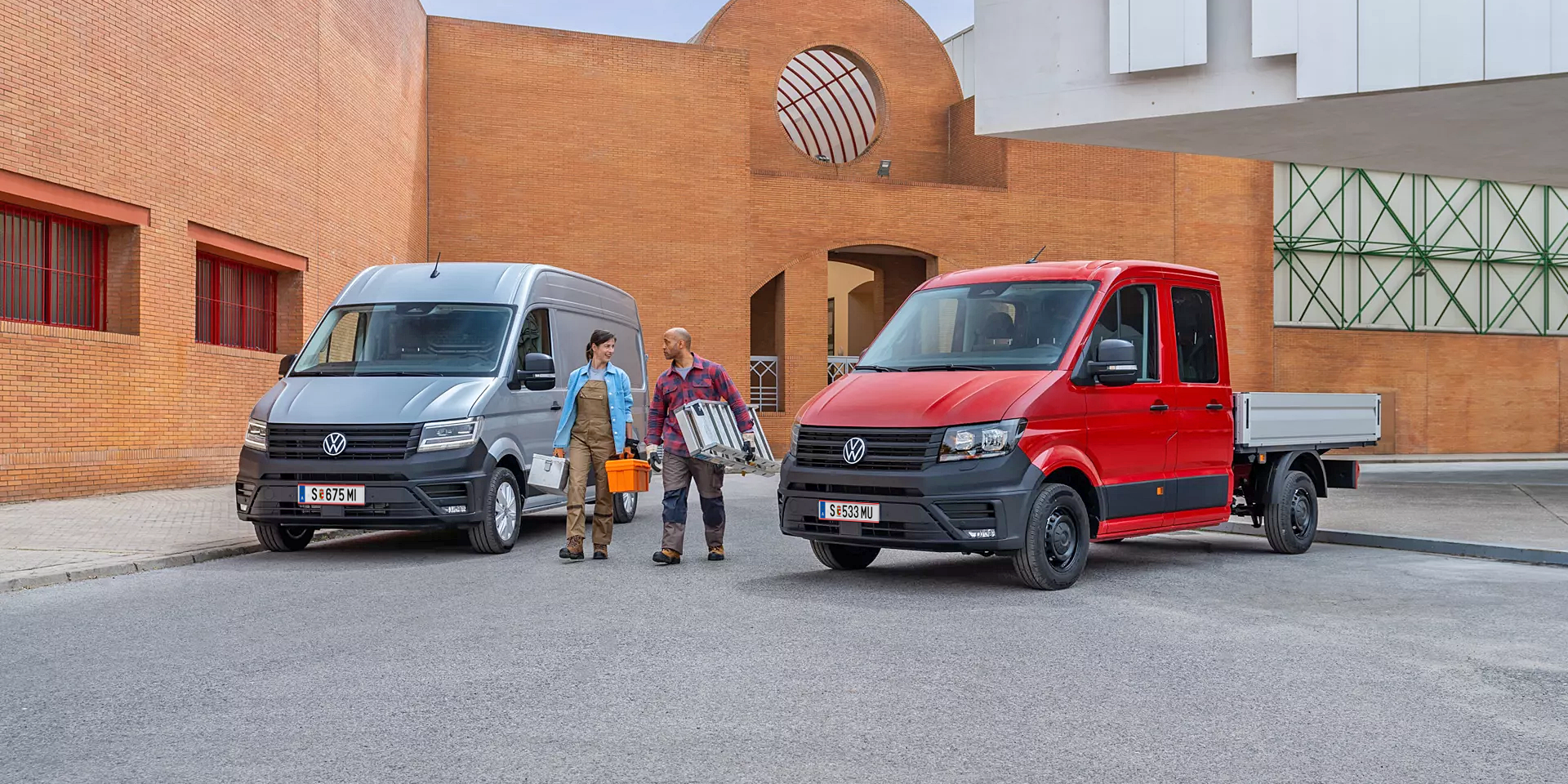 Два товарни автомобила Volkswagen Crafter – един закрит сив ван и един червен с открита платформа, паркирани на строителен обект с двама работници