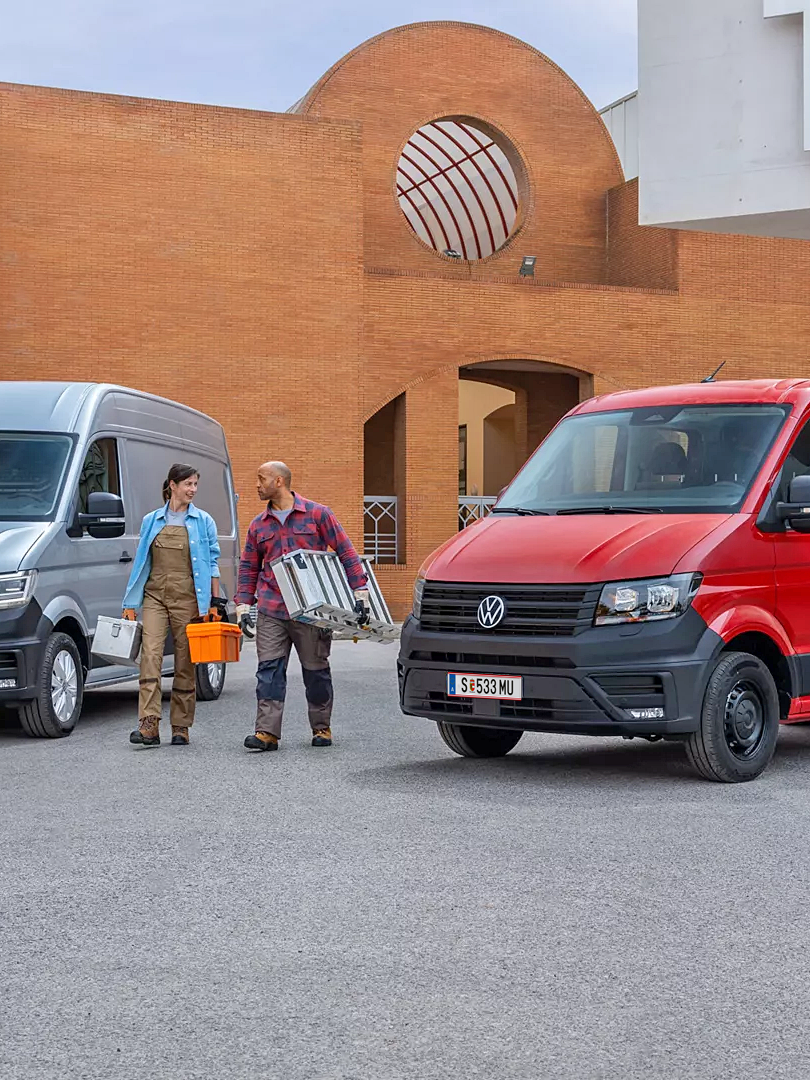 Два товарни автомобила Volkswagen Crafter – един закрит сив ван и един червен с открита платформа, паркирани на строителен обект с двама работници