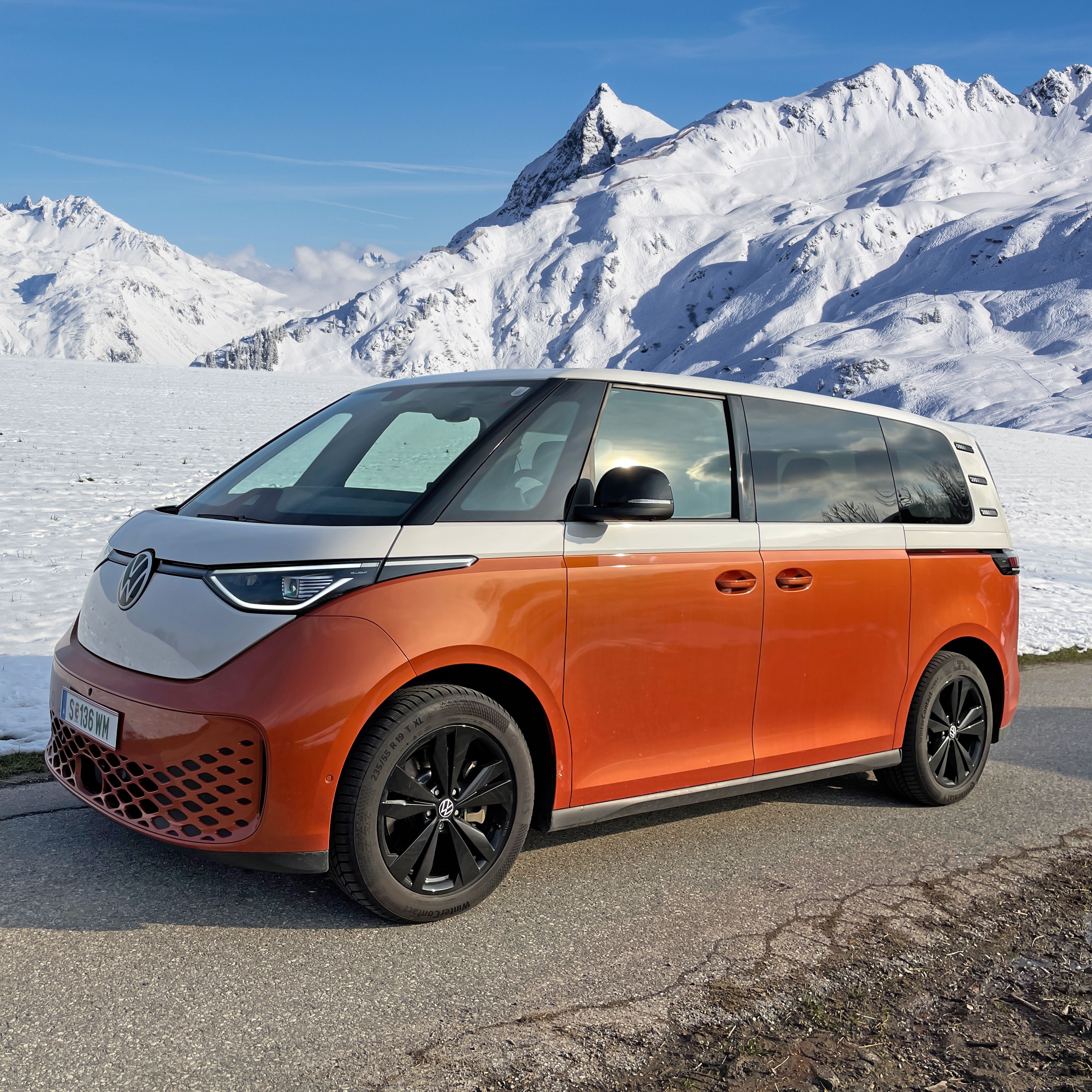 Електрически Volkswagen ID. Buzz в оранжево-бяла цветова комбинация на фона на заснежени алпийски планини.