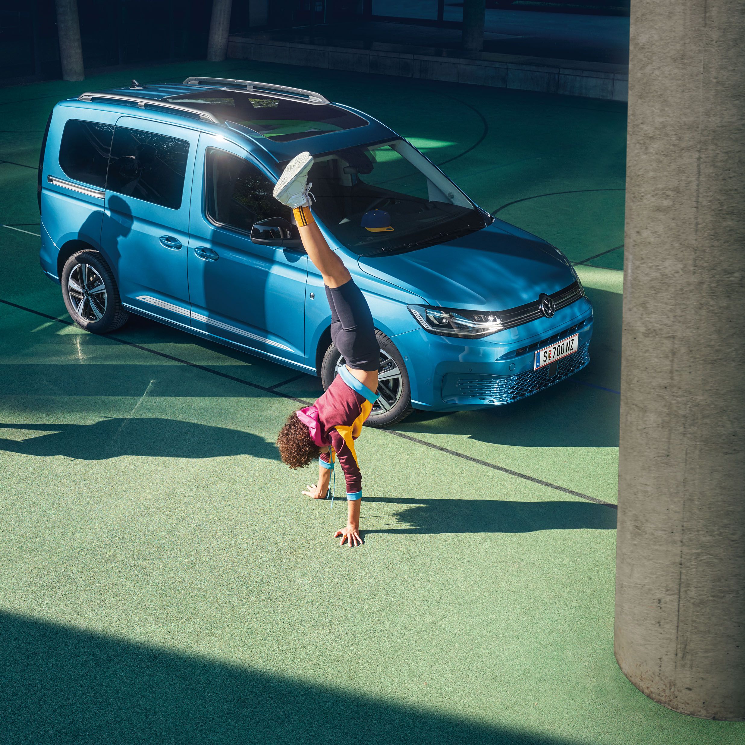Млади хора играят баскетбол и правят стойка на ръце пред син Volkswagen Caddy, паркиран на спортно игрище.