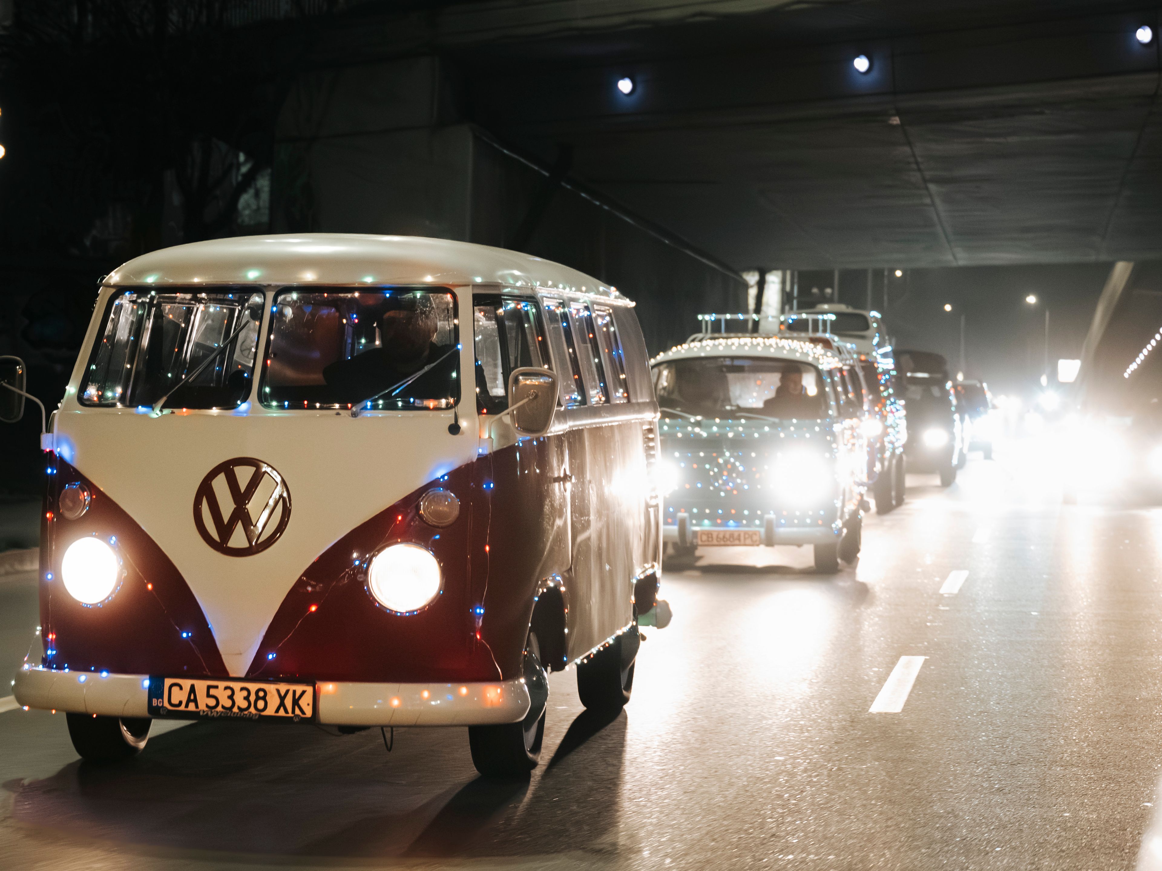 Две стари микробуса Volkswagen, украсени с празнични светлини, които се движат по нощна градска улица под мост. Вижда се регистрационният номер на първия микробус. Улицата е слабо осветена.