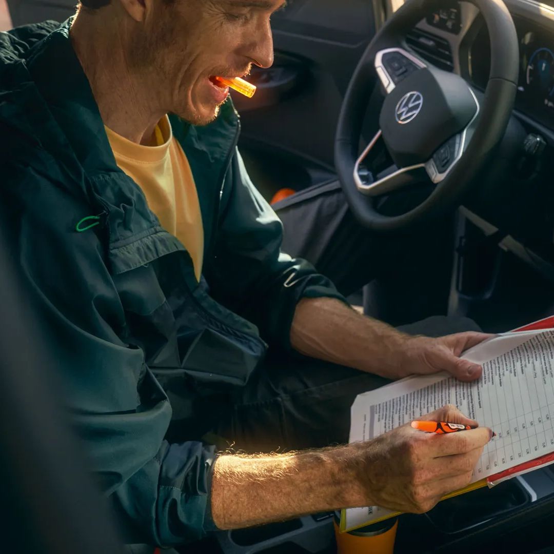 Човек попълва формуляр на клипборд с химикалка, докато седи в кола с навигационен екран, показващ карта на таблото.