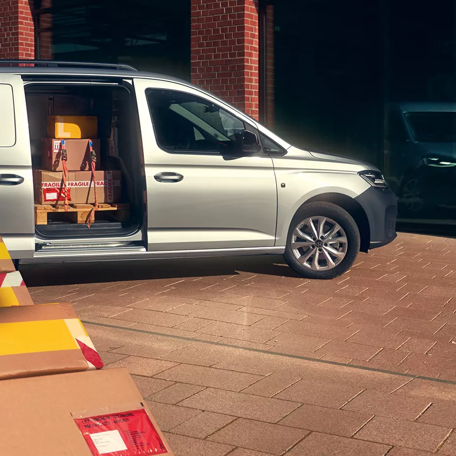 Куриер с количка пренася пакети пред сребрист товарен автомобил Volkswagen с отворена странична врата, натоварен с кашони.