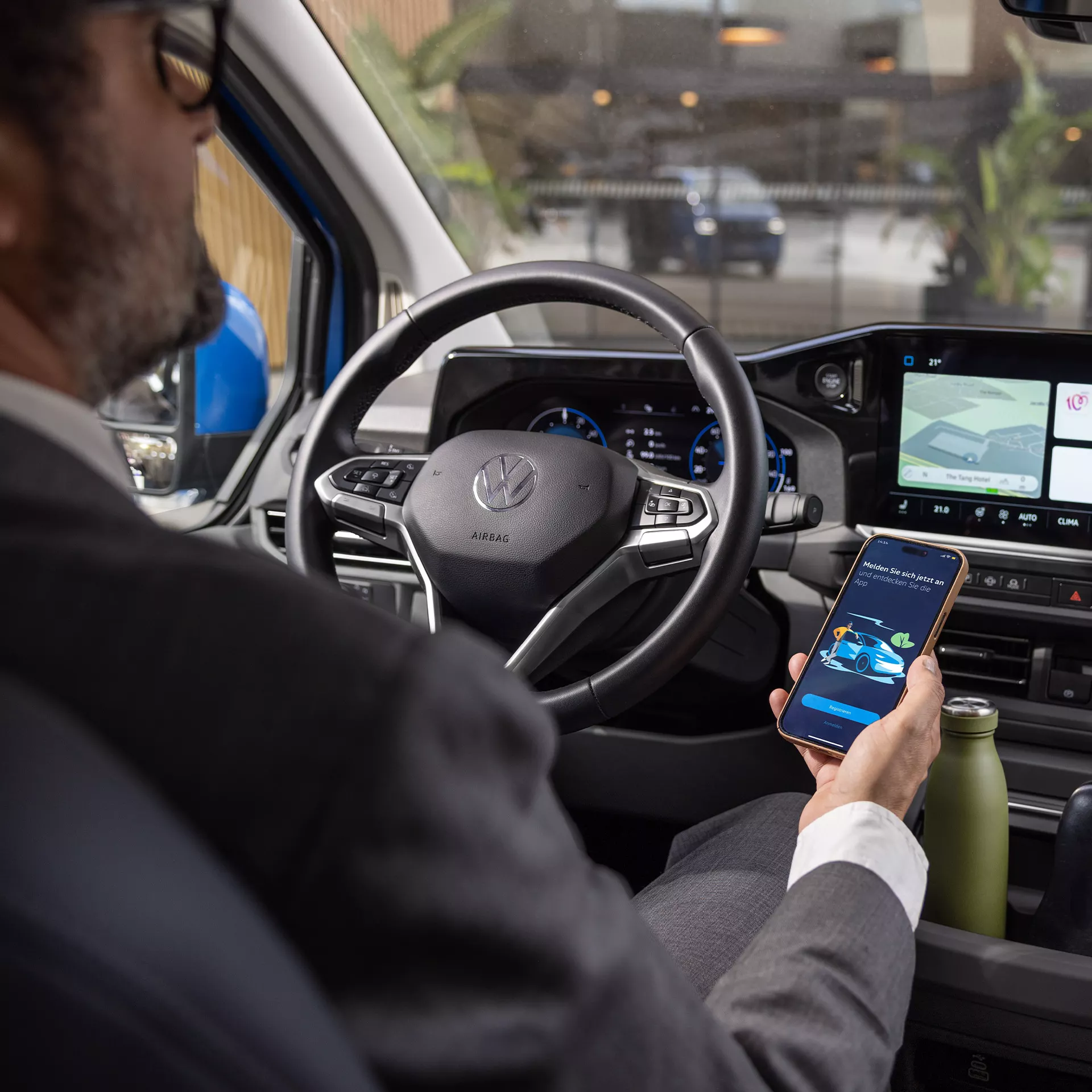 Мъж в костюм зад волана на Volkswagen Caravelle, използващ мобилно приложение за управление на функциите на автомобила.