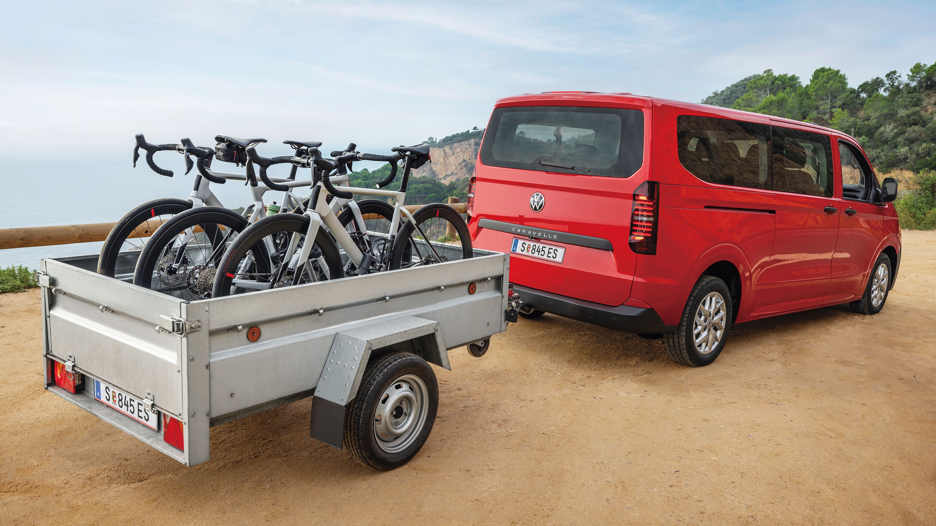 Volkswagen Caravelle в червено с ремарке и велосипеди, паркиран край живописен бряг
