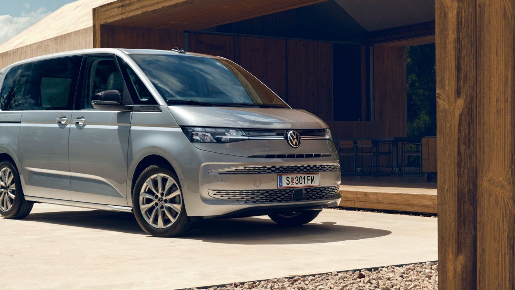 Двама мъже с багаж до сребрист Volkswagen Multivan, паркиран пред модерна къща.