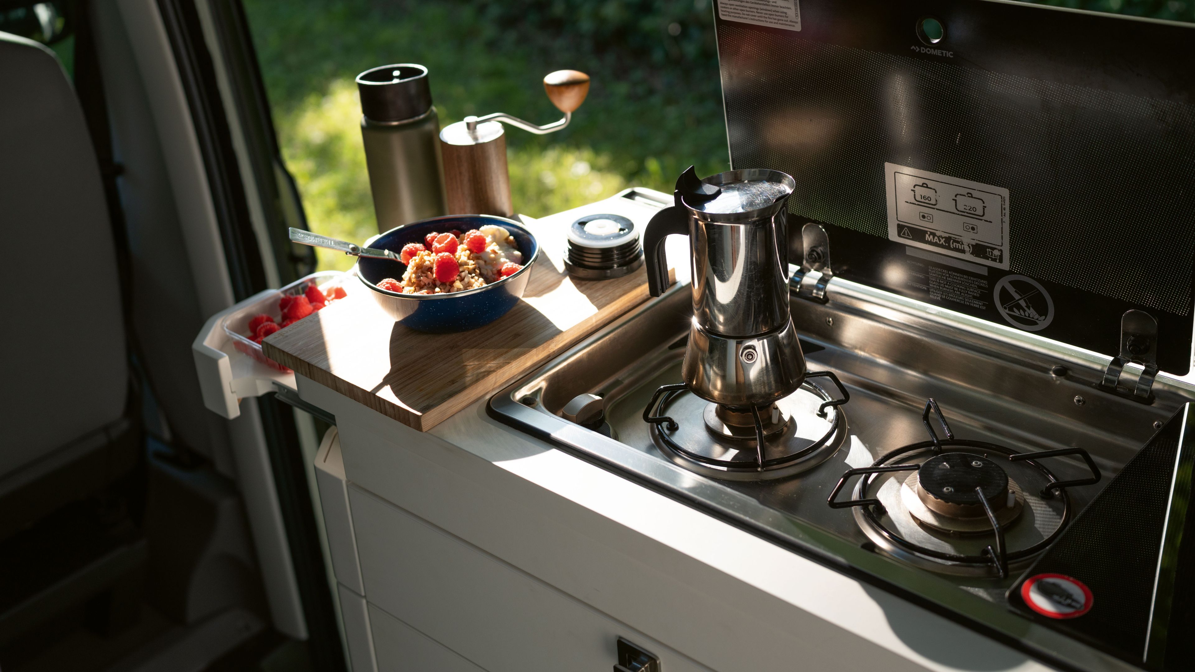 Ein Campervan mit einer eingebauten Küche ist abgebildet. Auf der Arbeitsplatte stehen eine Kaffeekanne, eine Müslischale und eine Thermoskanne. Im Hintergrund ist eine Grünfläche zu sehen.