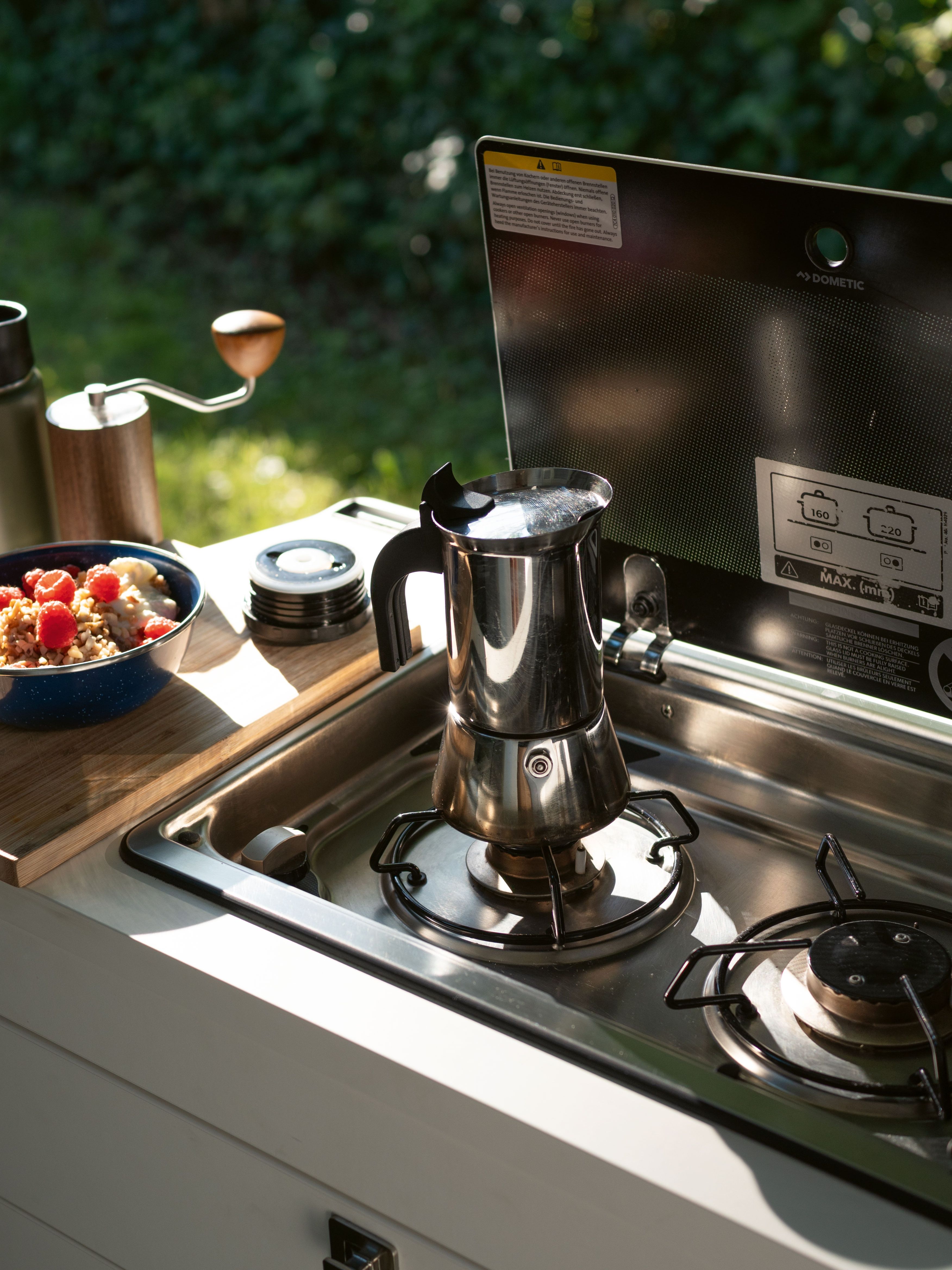 Ein Campervan mit einer eingebauten Küche ist abgebildet. Auf der Arbeitsplatte stehen eine Kaffeekanne, eine Müslischale und eine Thermoskanne. Im Hintergrund ist eine Grünfläche zu sehen.