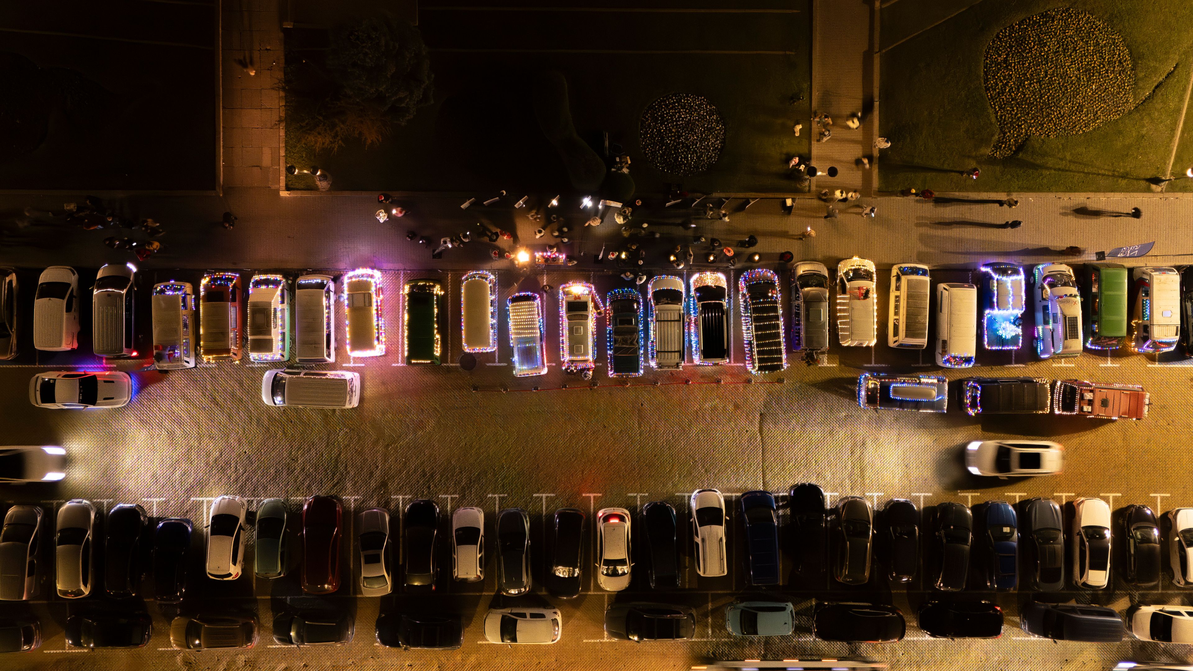 Паркирани Volkswagen автомобили под улично осветление, снимани от птичи поглед. В горната част на изображението има лица и обекти, осветени от полицейски светлини. Пешаходна зона и зелена площ са видими също на изображението.