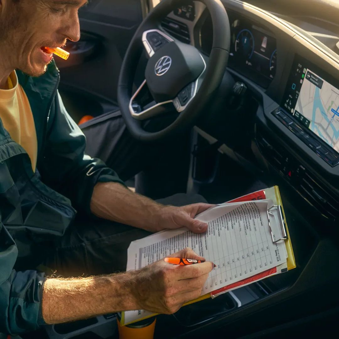 Човек попълва формуляр на клипборд с химикалка, докато седи в кола с навигационен екран, показващ карта на таблото.