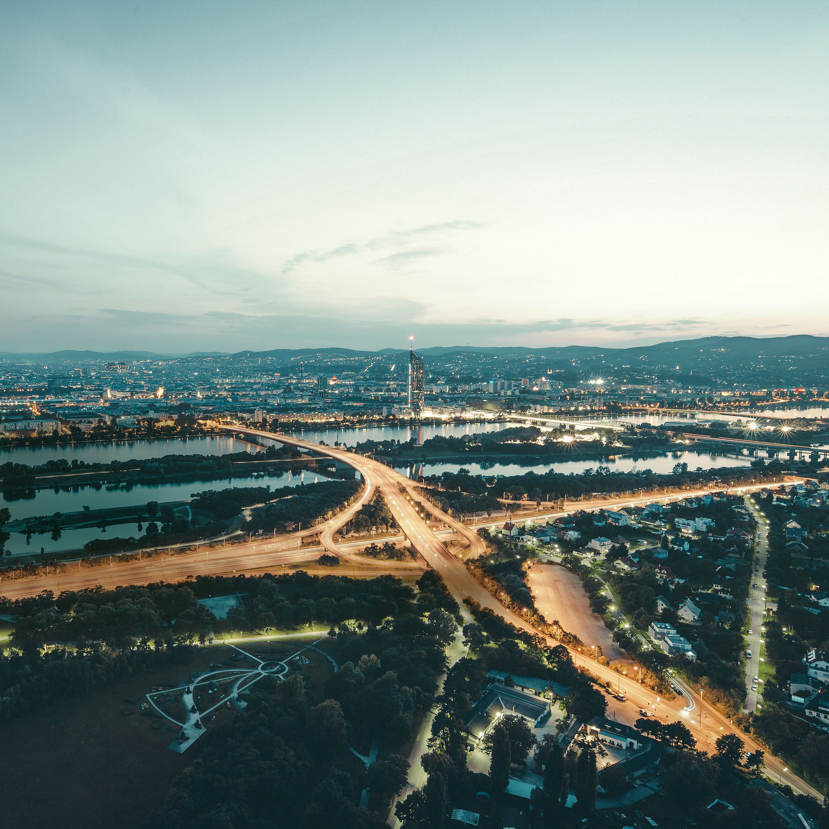 Панорамна снимка на голям град с река и мостове. Градът е осветен по време на залез. На преден план има квартал с жилища и зелени площи.