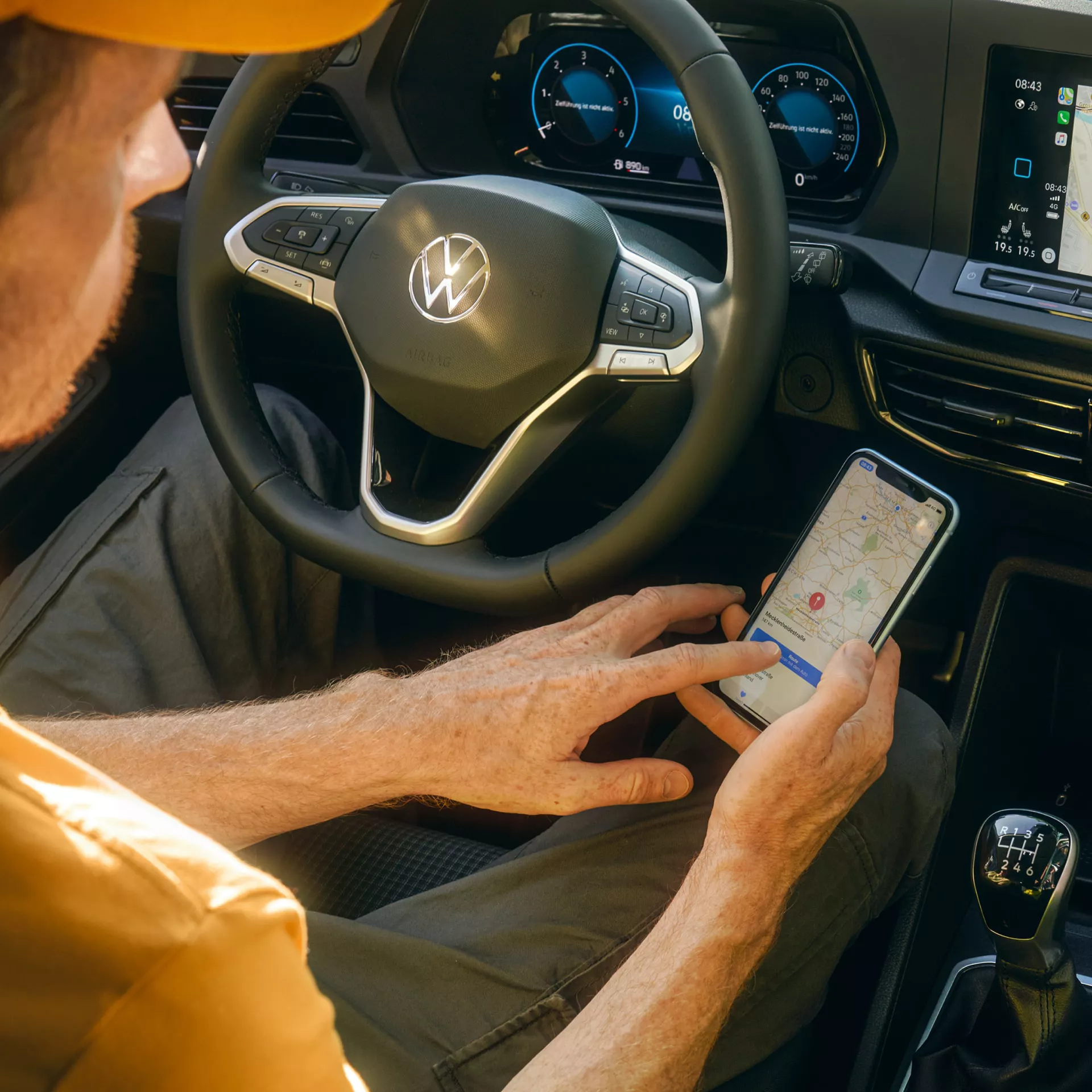 Куриер използва навигационно приложение на смартфон, седнал зад волана на автомобил Volkswagen с дигитален кокпит и модерен мултимедиен екран.