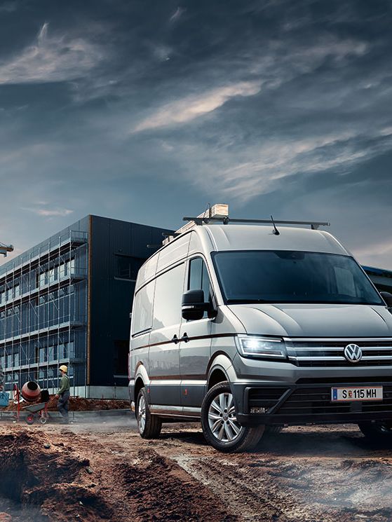 лекотоварен автомобил Volkswagen е паркиран пред строеж. В близост до него се виждат двама строителни работници. На заден план има сглобяема сграда и двата строителни крана.