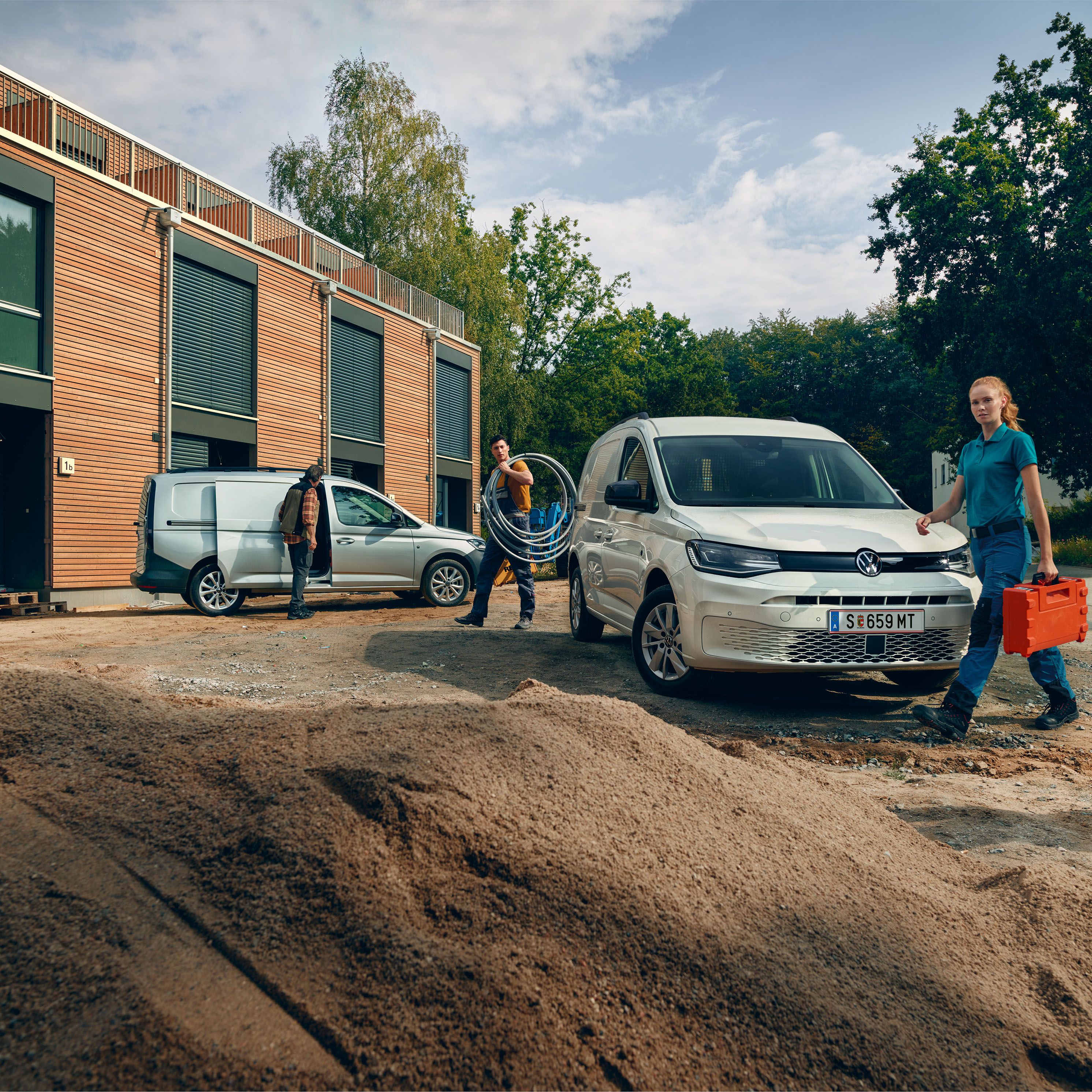 Два товарни автомобила Volkswagen Caddy на строителен обект, с трима професионалисти, носещи инструменти и оборудване.